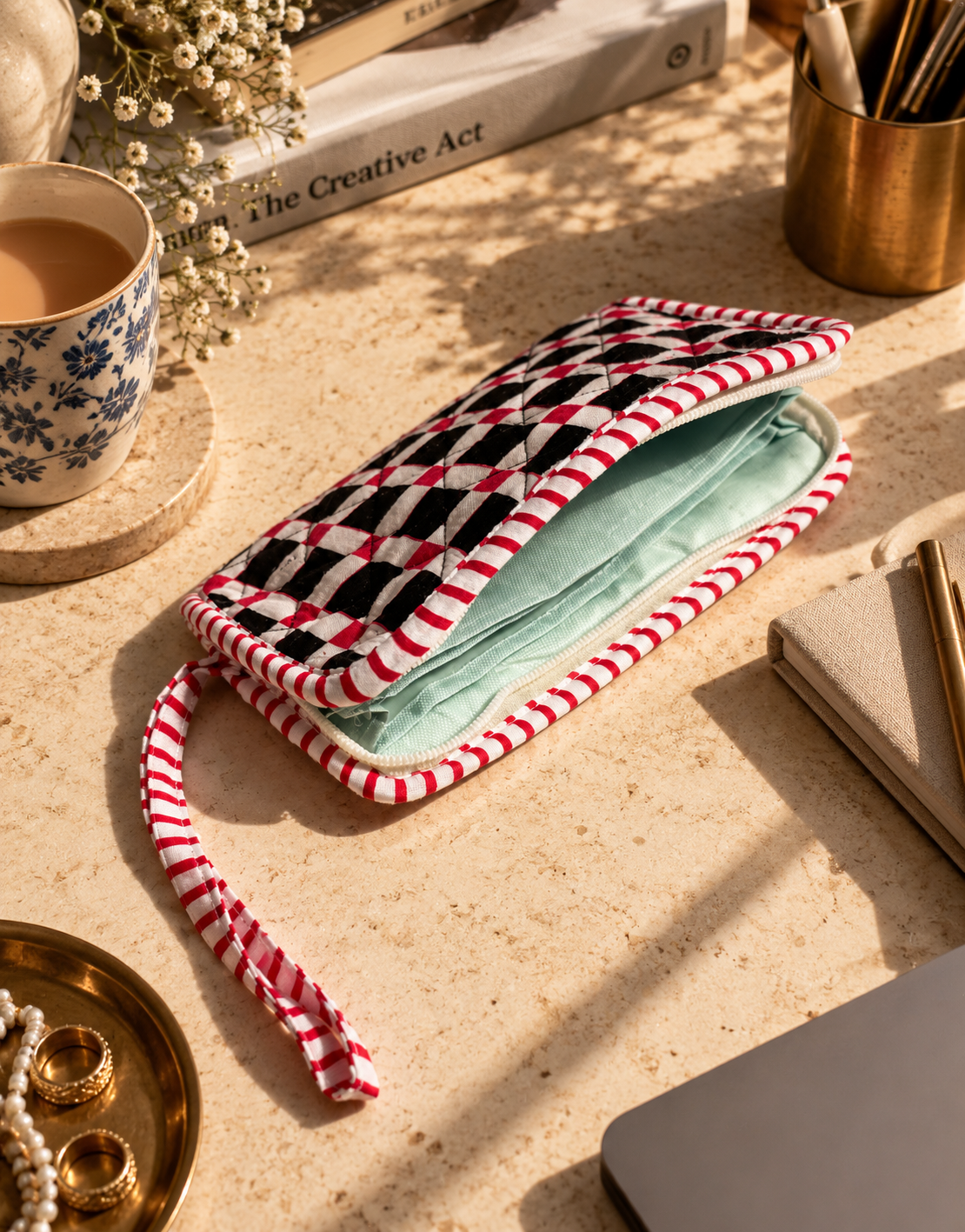 A black & white mixed with red cotton quilted wallet/clutch with its zipper opened showcasing the water-resistant interiors with sections on a table with other accessories and a cup of tea.
