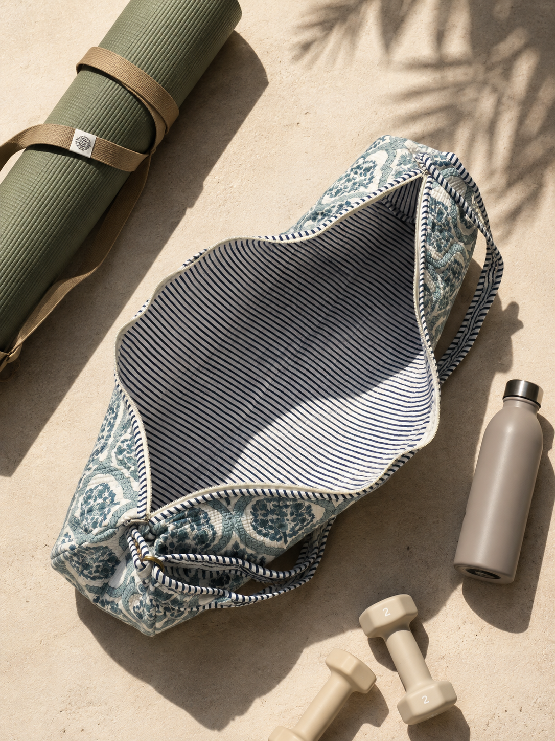 A patterned yoga mat bag with a blue and white paisley print and striped interior, styled on a sandy surface with a rolled yoga mat, dumbbells, and a water bottle nearby.