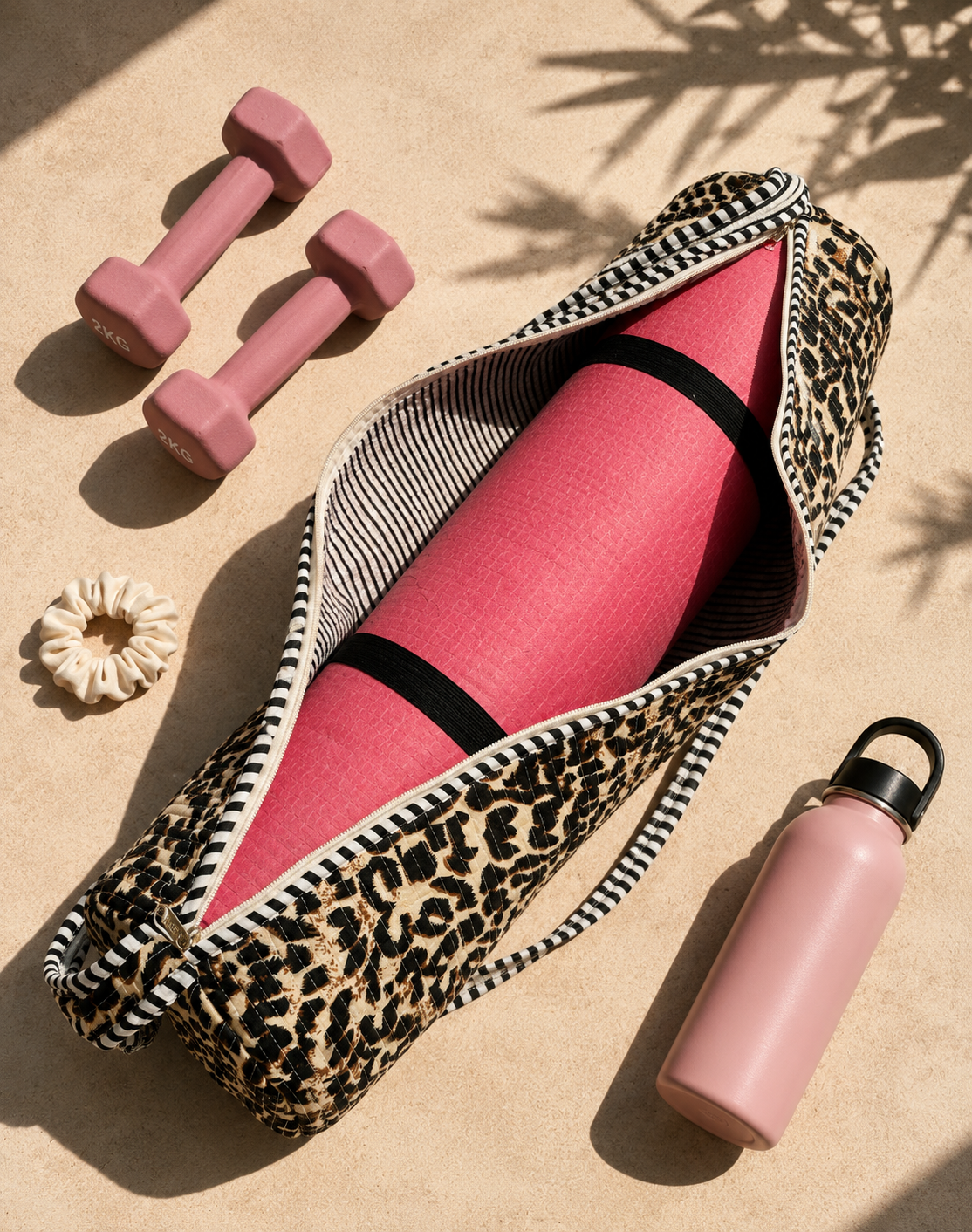 A leopard-print zippered gym bag lies open on a sandy surface, containing a rolled pink yoga mat with black bands. Surrounding props include two pink dumbbells, a pink water bottle, and a cream scrunchie under dappled sunlight.
