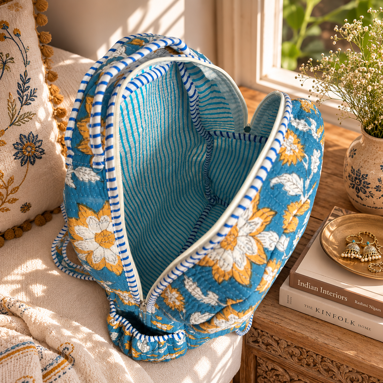 A blue quilted fabric backpack with a floral pattern, striped trim blue and white striped interiors, styled on a white armchair in a warm, sunlit room next to a wooden table with books and flowers.
