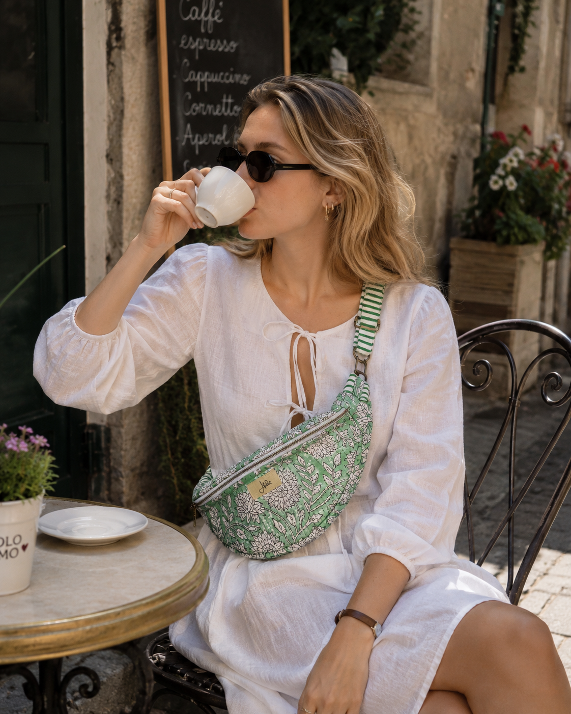 A woman in a white dress sits at an outdoor cafe table drinking from a white mug, wearing a green and white patterned floral crossbody fanny bag with a striped shoulder strap.