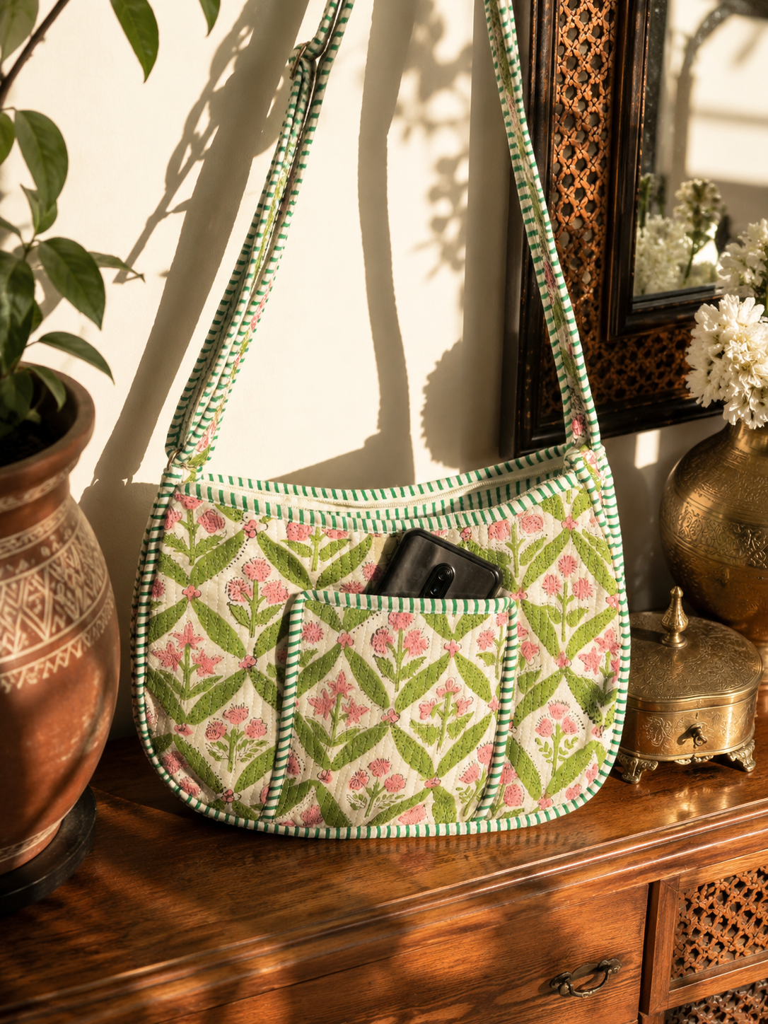 A green and pink floral embroidered shoulder bag with a striped trim, resting on a wooden cabinet next to a potted plant and brass decorative items.