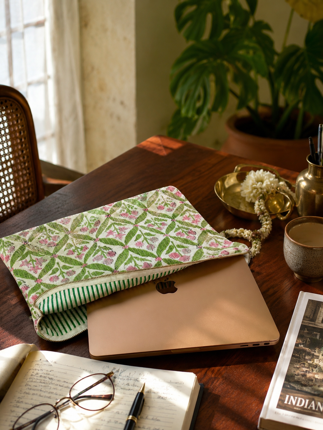 A green and pink floral quilted laptop sleeve with a green and white striped interior holding a laptop on a wooden study table with a notebook, glasses, pen and flowers.