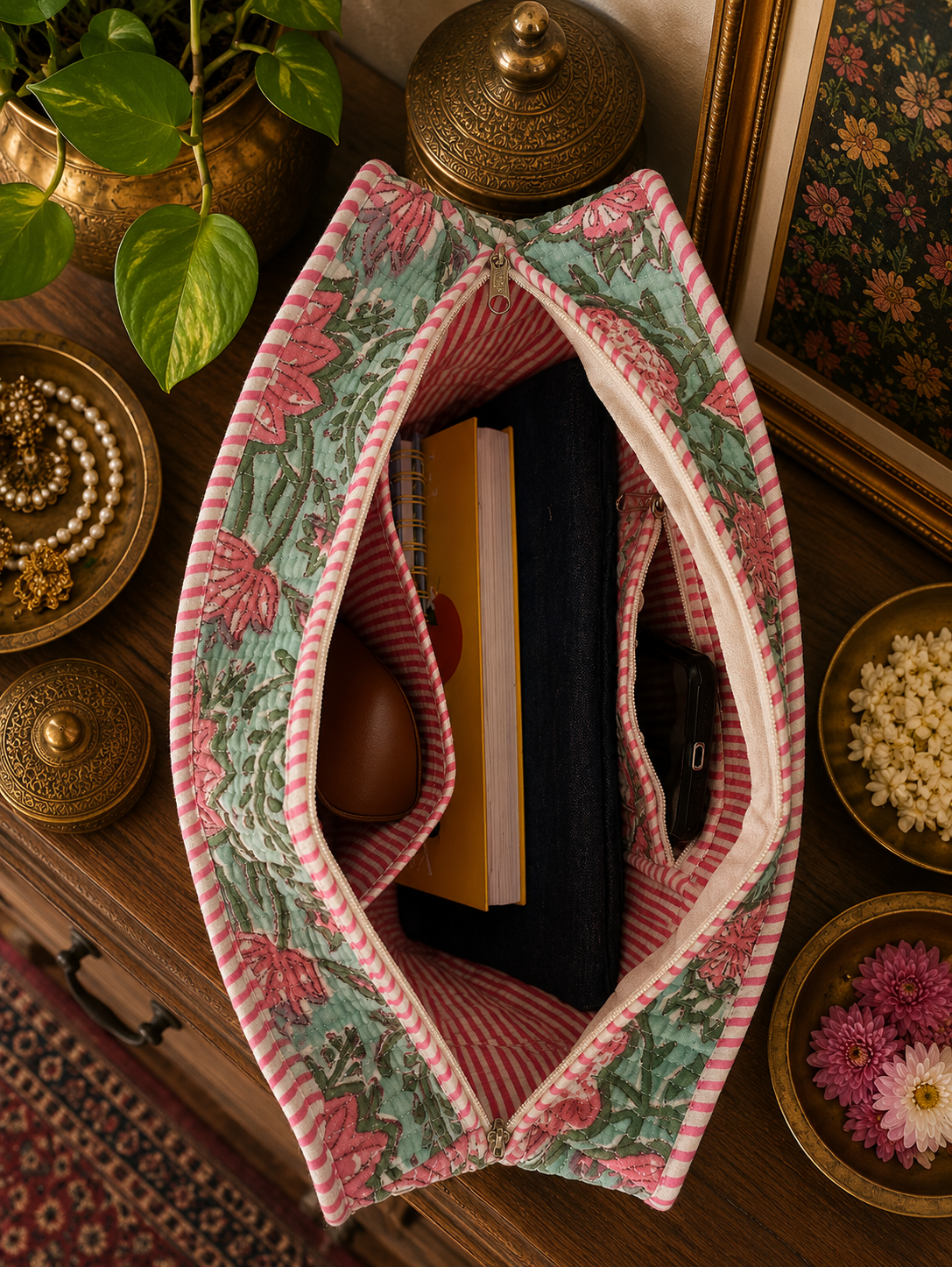A floral-patterned quilted zipper tote bag on a wooden surface with decorative items around. The bag is open and contains a laptop sleeve, notepads, sunglass cover and a mobile phone.