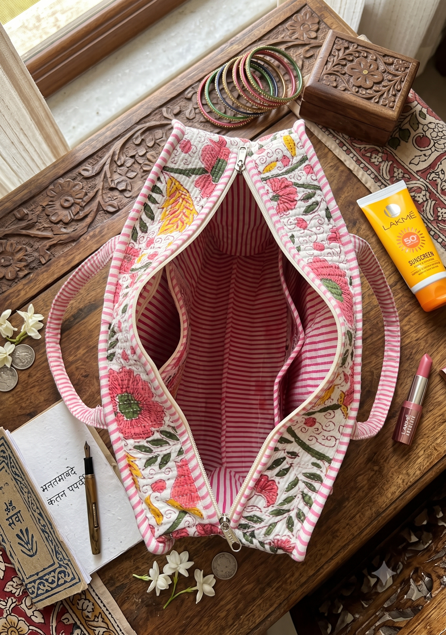 A pink and ywellow floral quilted tote bag with pink and white striped interiors laid top facing on a carved wooden table, surrounded by small lifestyle props including bangles, a tube of body cream, lipstick, coins, and a notebook. The inside of the bag has 3 pockets.