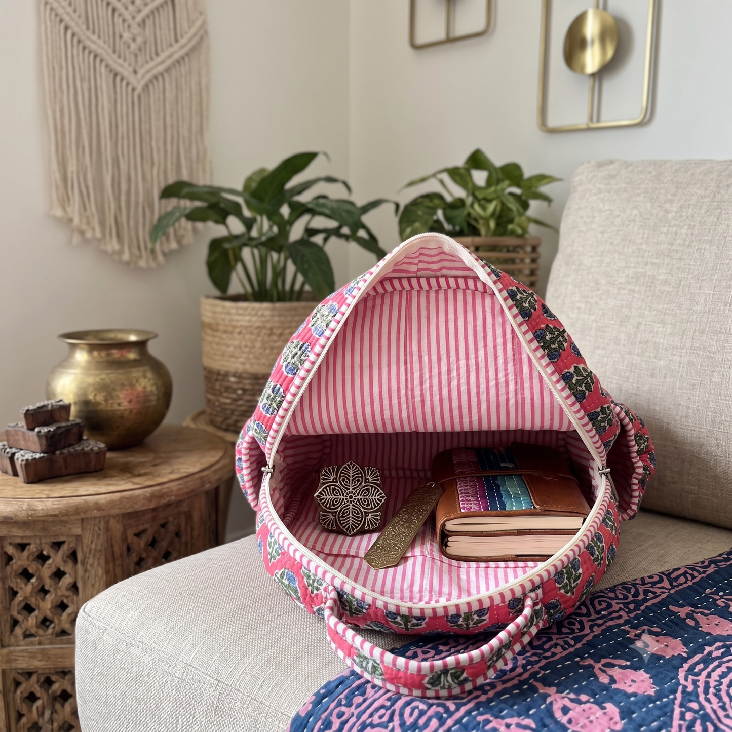 A pink and multicolor floral backpack with a striped interior lining sits open on a light beige sofa, styled with a book and small decorative item inside. The background shows a cozy living room setting with potted plants, a brass pot on a wooden side table, and wall decor.