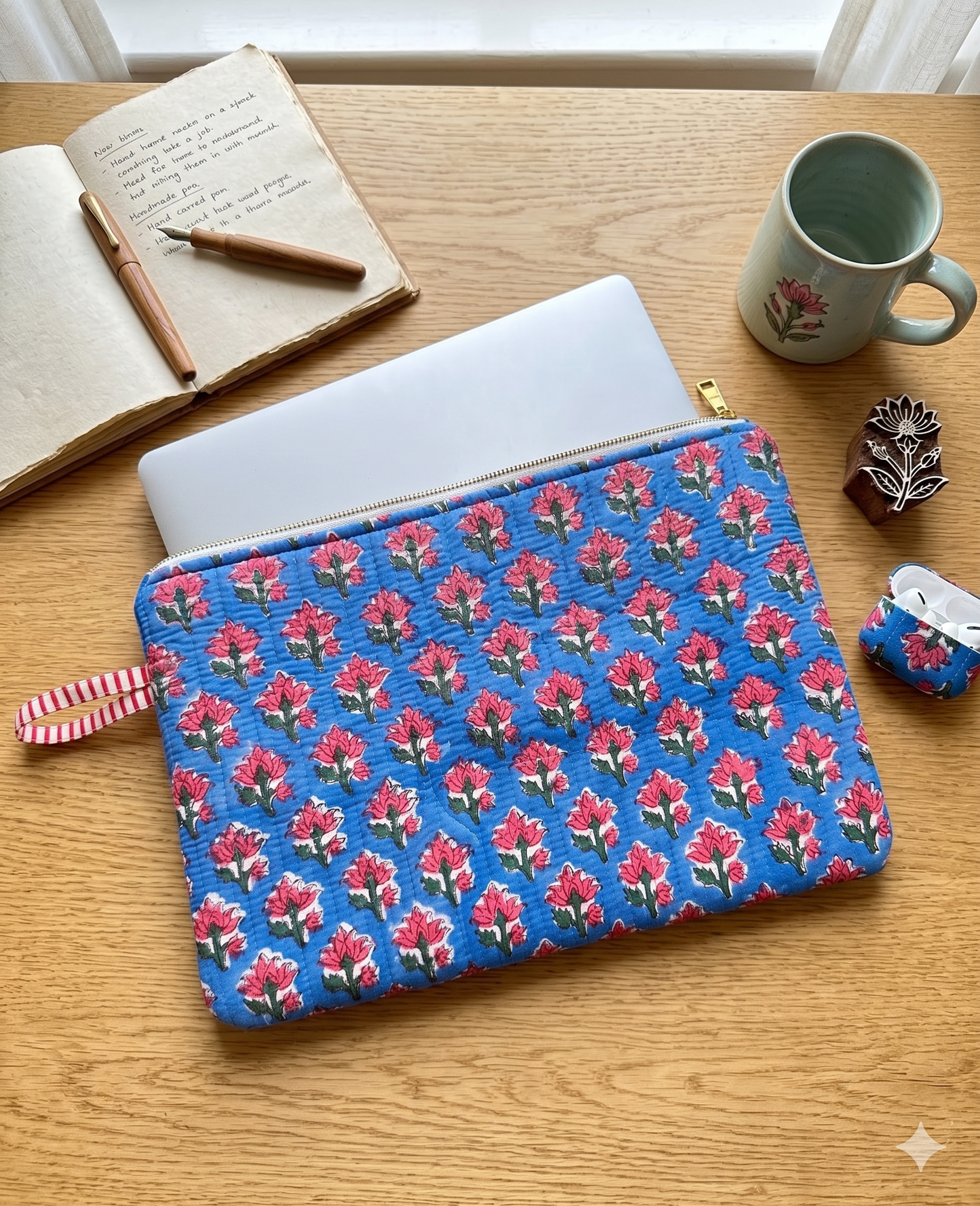 A blue quilted laptop sleeve with a pink and green floral blockprint pattern all over, placed on a wooden tabletop alongside a coffee mug, a wooden block, an AirPod case, and a notebook with a pen.