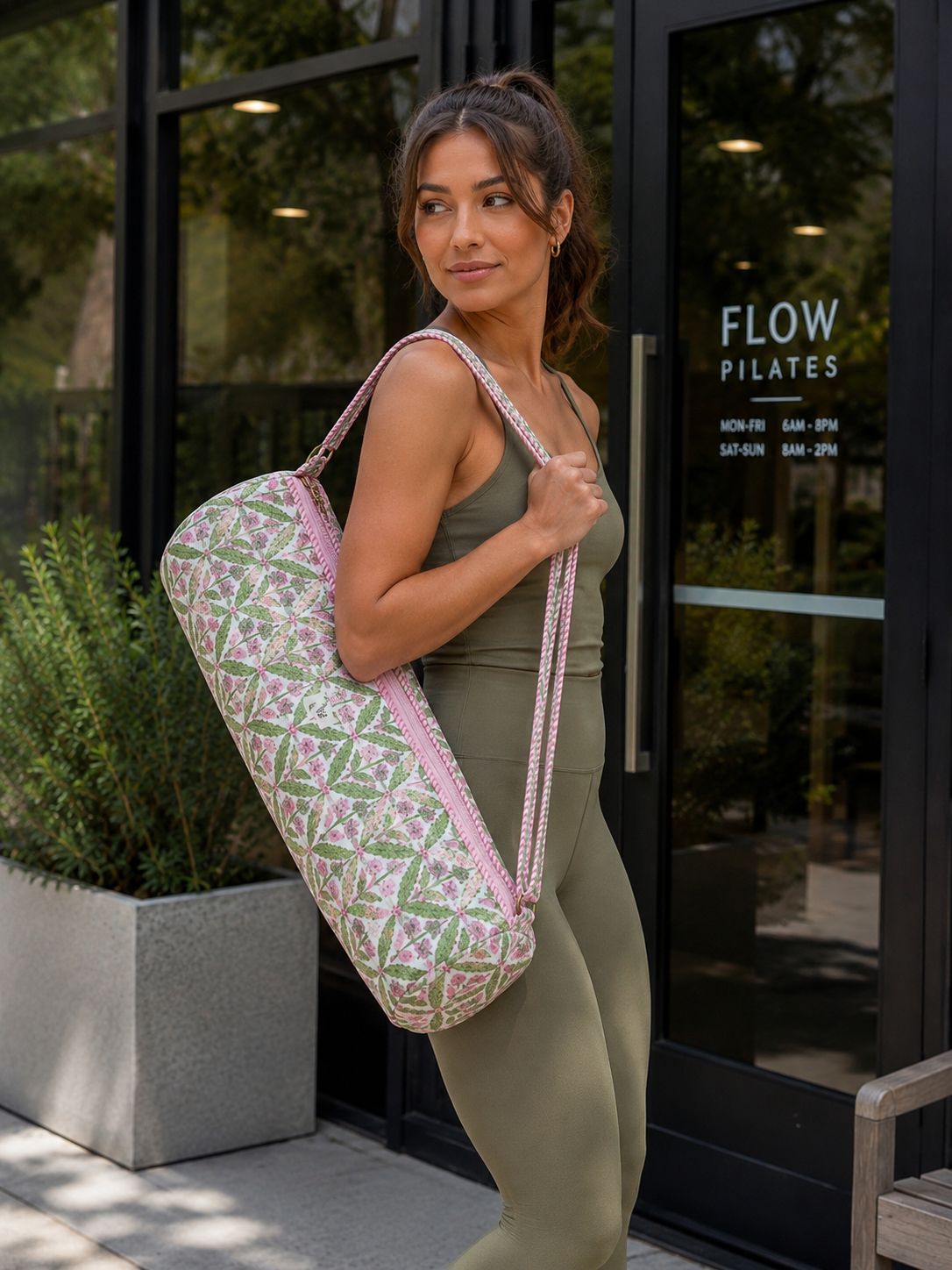 A woman in olive green activewear carries a large, patterned yoga mat cover bag with pink straps and a geometric print, standing outside a fitness studio with a glass door labeled 'FLOW PILATES'.