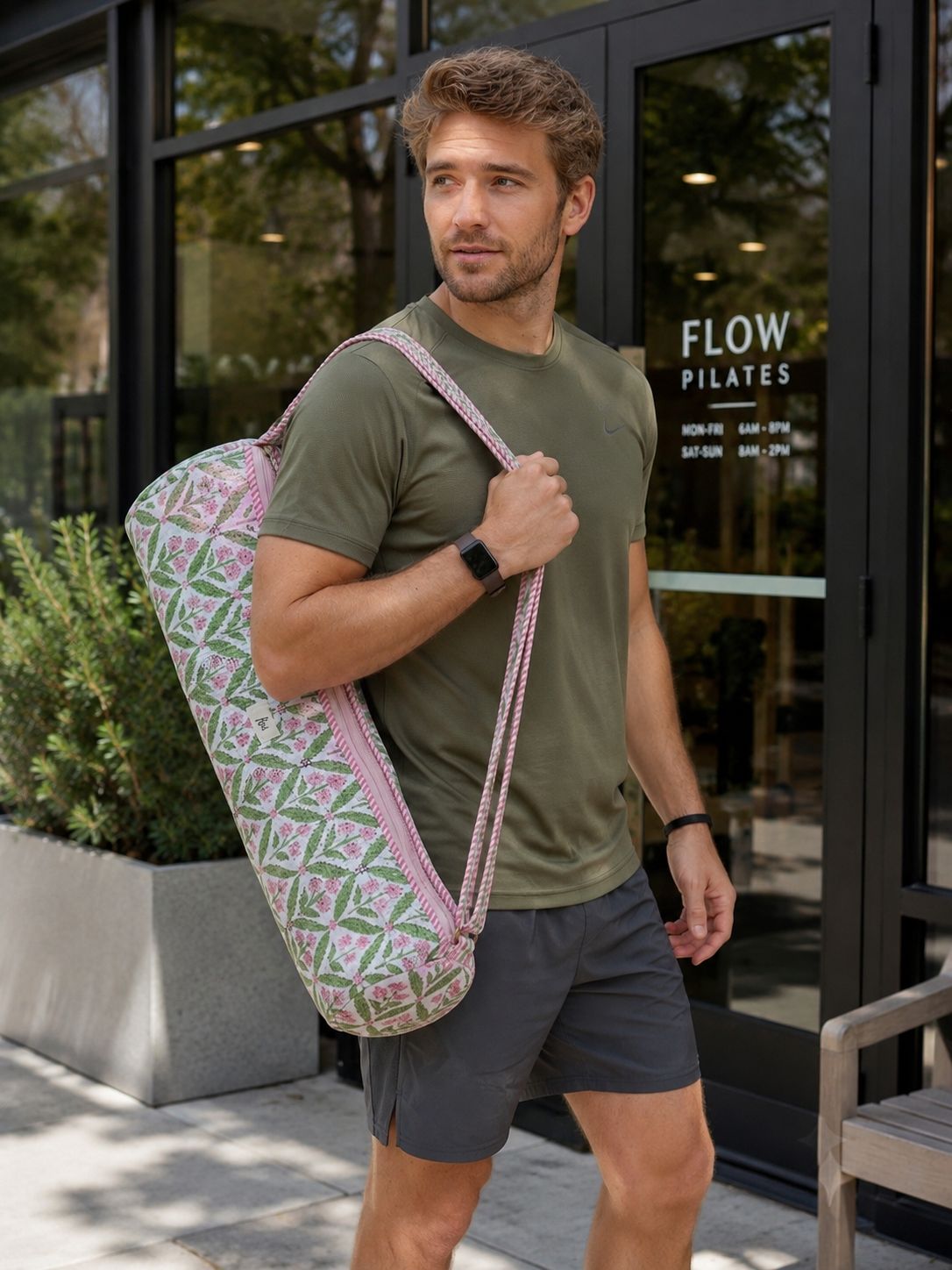 A man wearing an olive green t-shirt and gray shorts carries a pink and green patterned yoga mat cover bag over his shoulder as he walks outside a building with 'FLOW PILATES' written on the glass door.