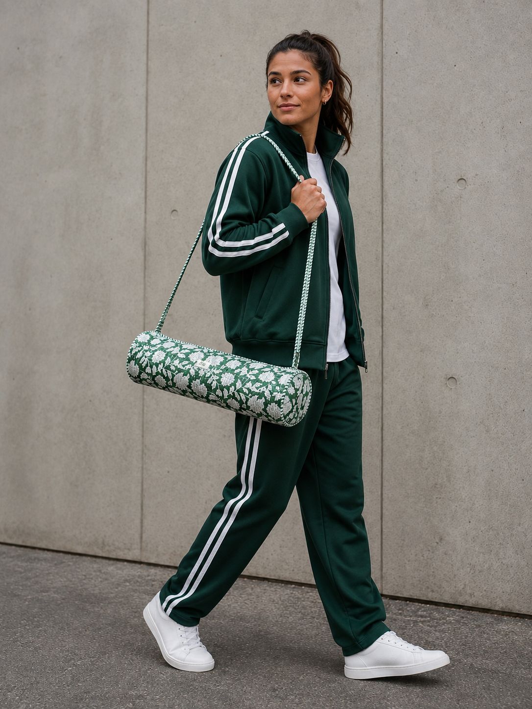 A woman walking outdoors wearing a dark green athletic tracksuit with white stripes, a white t-shirt, and white sneakers, while carrying a green and white floral patterned quilted yoga mat cover bag.