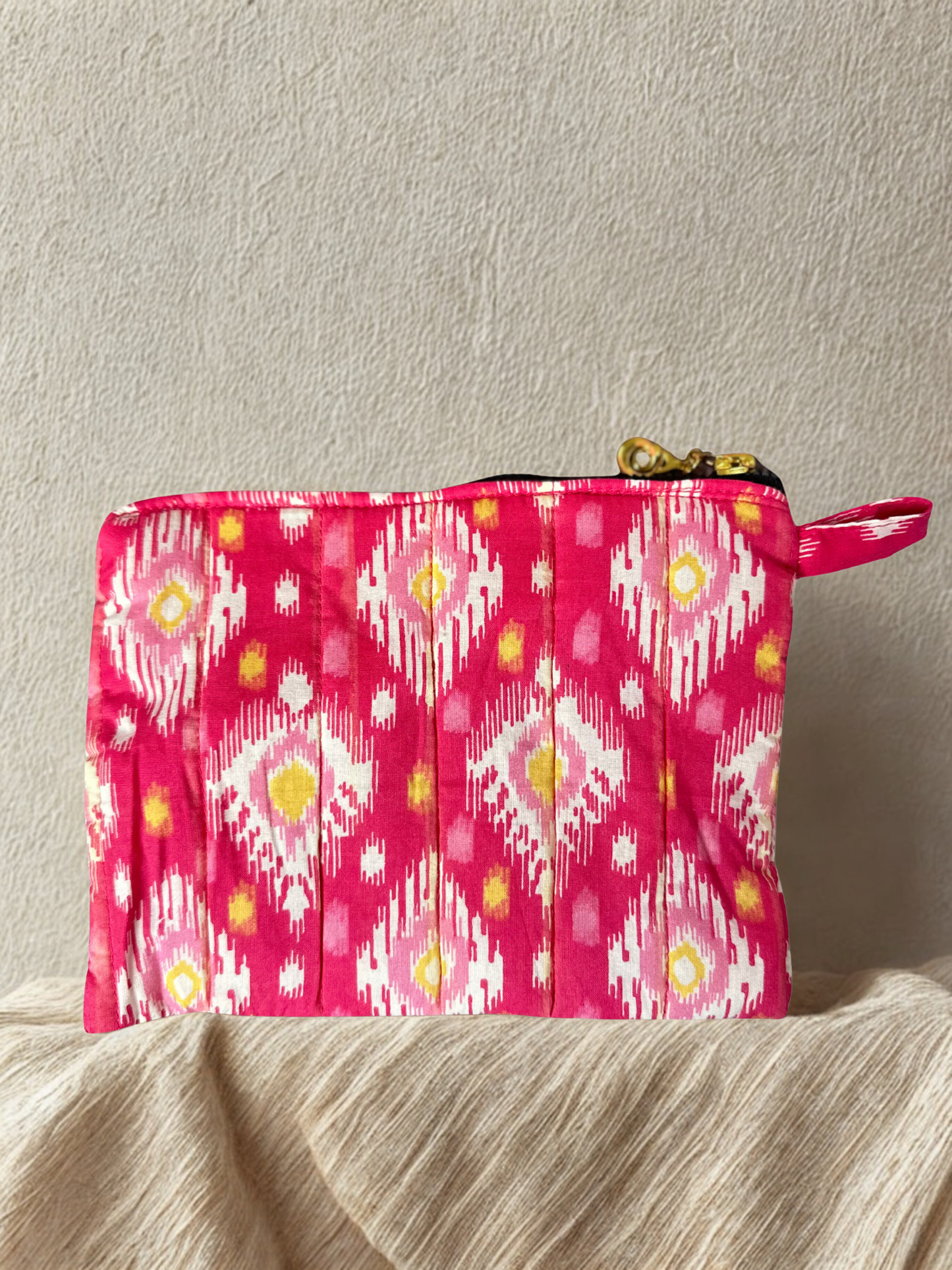 A pink patterned zippered pouch on an earthy tone linen and beige wall
