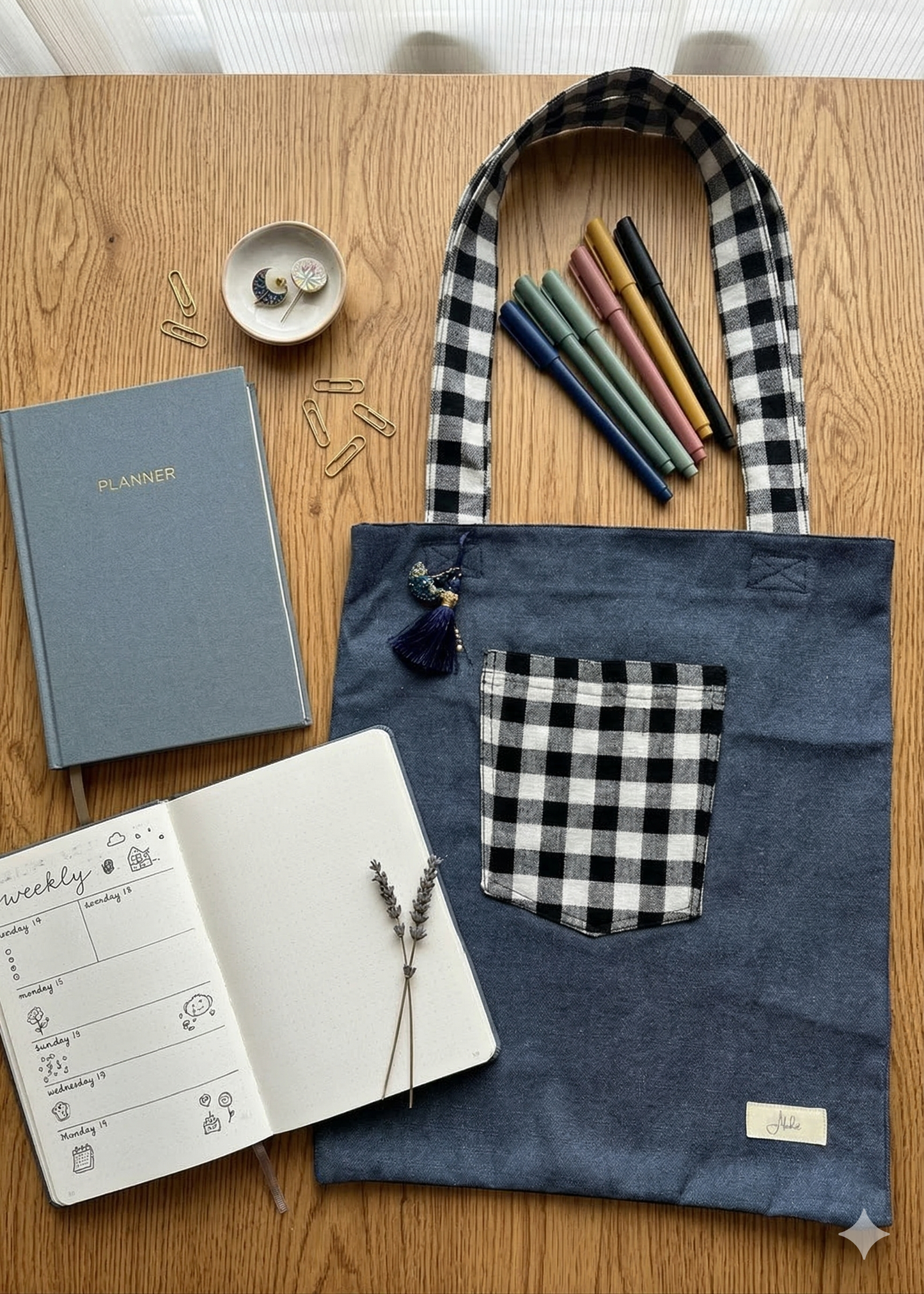 A greyish-blue denim tote bag with plaid black & white front pocket and straps on a wooden surface with stationery items.