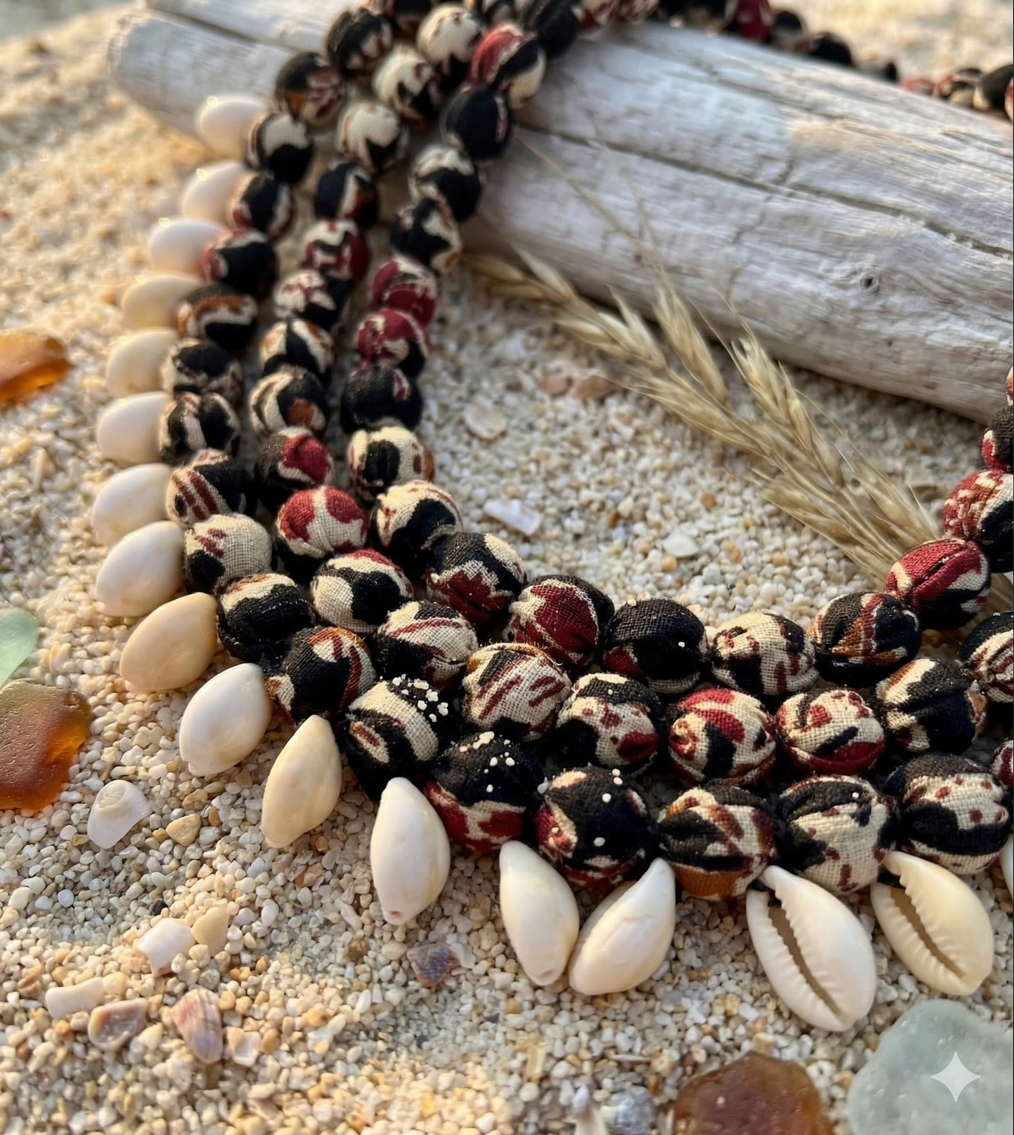 Handmade Layered fabric Necklace with  cowrie shells at the bottom layer on a sandy surface with driftwood and grass.