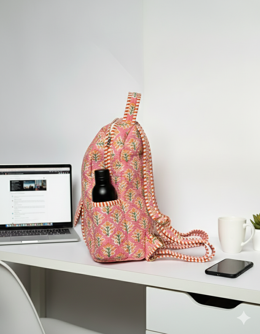 Pink floral quilted backpack with a bottle in teh side pocket on a white desk with a laptop and other items