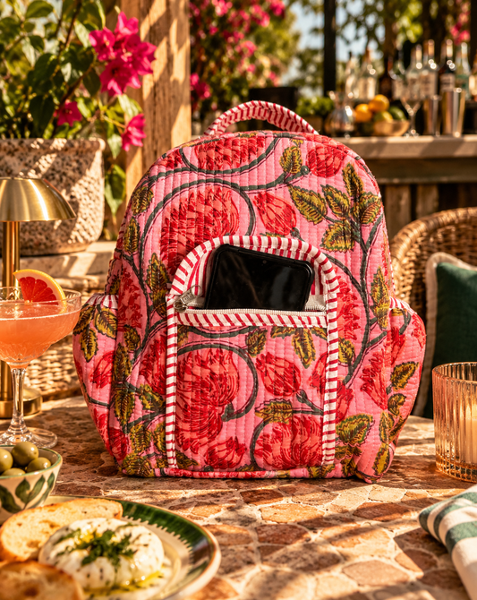 A pink floral backpack with red flowers and green leaves, featuring a striped zippered front pocket containing a black smartphone, set on a stone patio table with a cocktail, bread, and food in a warm outdoor dining environment.