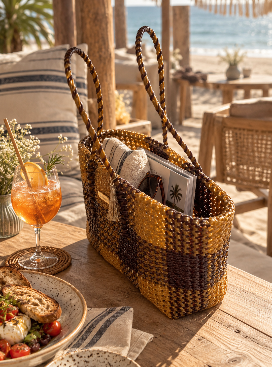 A woven beach tote bag in golden yellow, and brown stripes sits on a wooden table at a beachside cafe, styled with a drink, salad, and sunglasses.