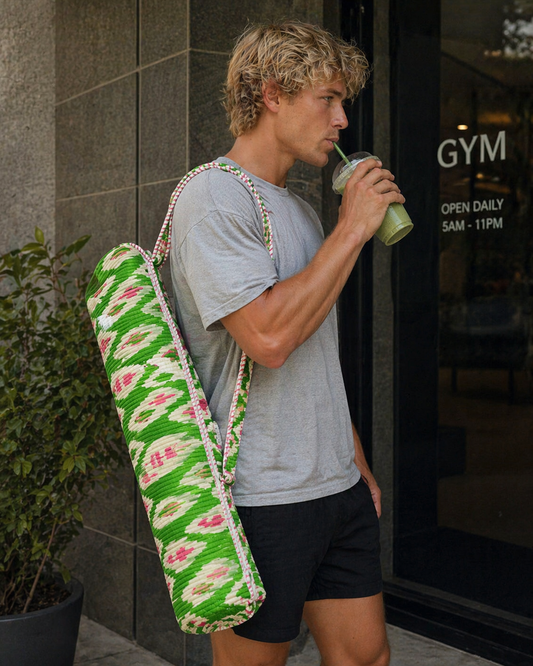 A man with a colourful green yoga mat bag and drink, standing outside a gym.
