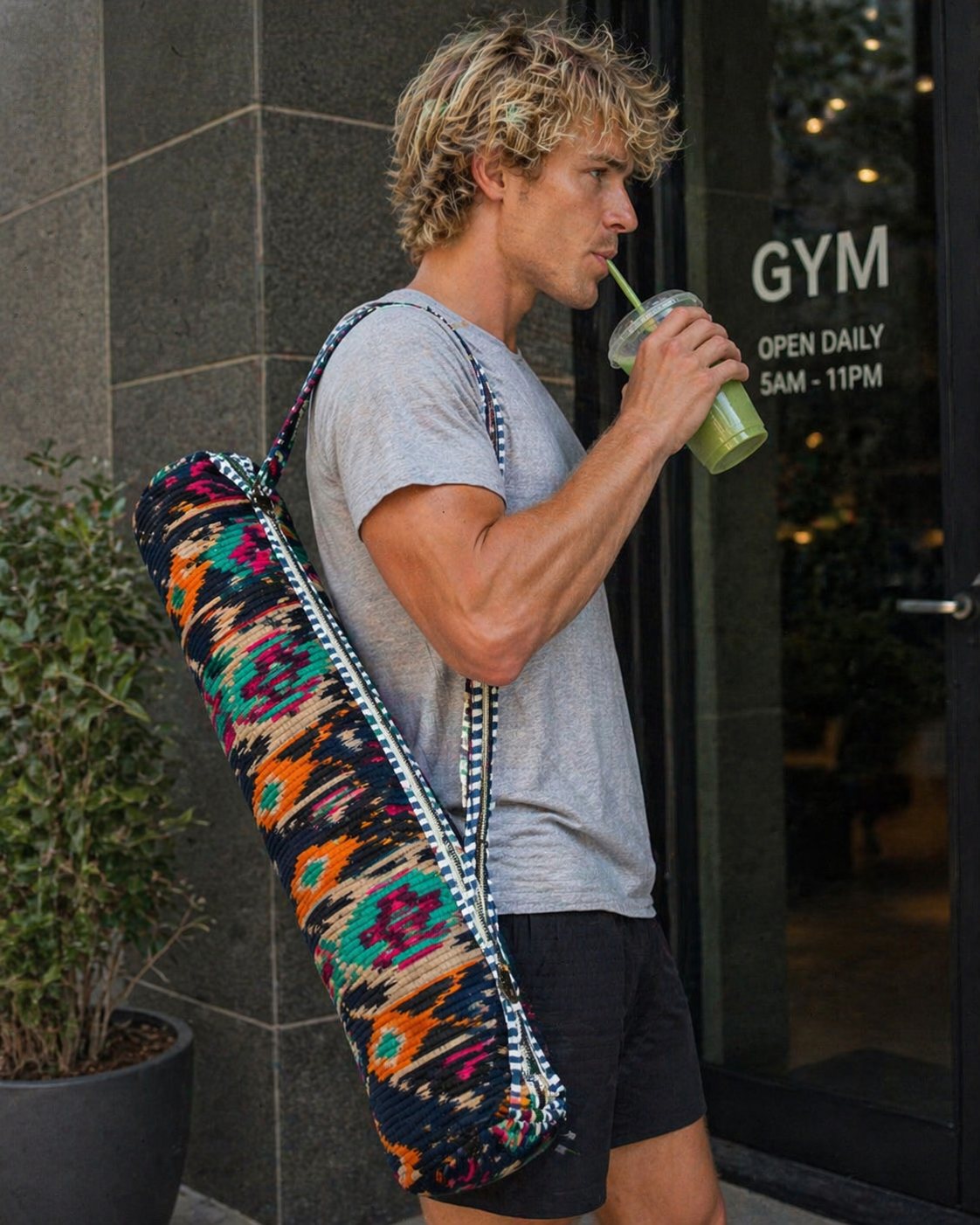 A man with a colourful ikat-print yoga mat bag drinks outside a gym.