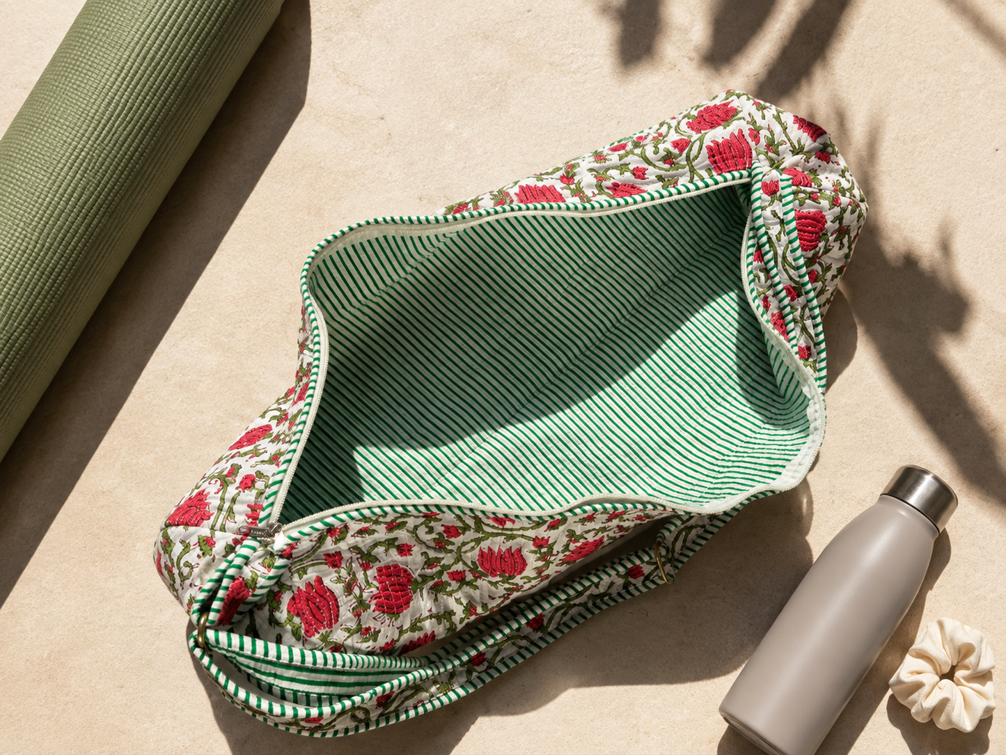 A fabric yoga mat bag with a green and white striped interior and a pink floral embroidered exterior is shown on a beige stone surface next to a rolled green yoga mat and a grey water bottle.