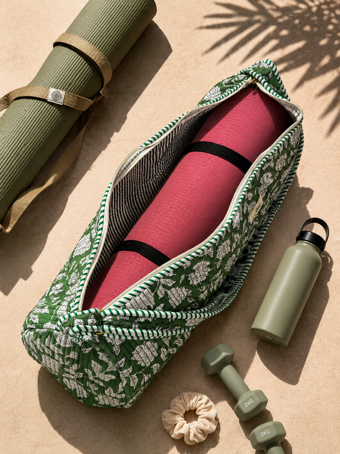 A green and white floral patterned yoga mat bag is shown open, containing a pink yoga mat. The scene is styled on a sandy surface with matching olive green fitness accessories, including a rolled yoga mat, a water bottle, dumbbells, and a scrunchie.
