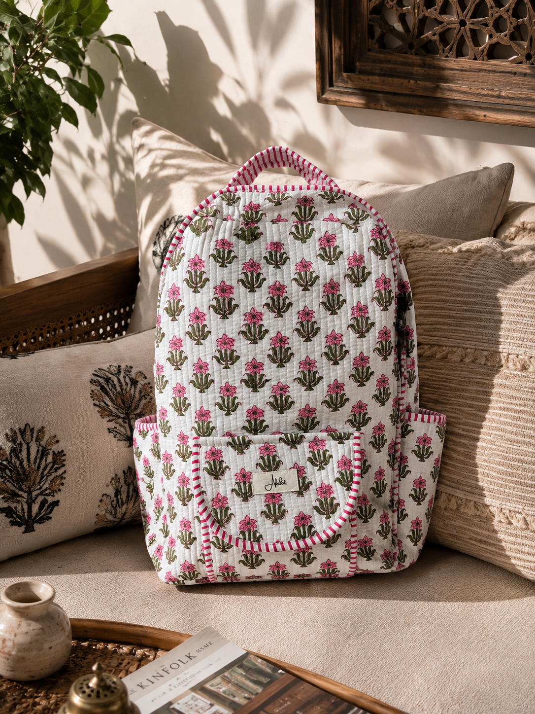 A quilted canvas backpack with a pink and green floral block print pattern, styled on a beige sofa with decorative pillows and warm natural sunlight.