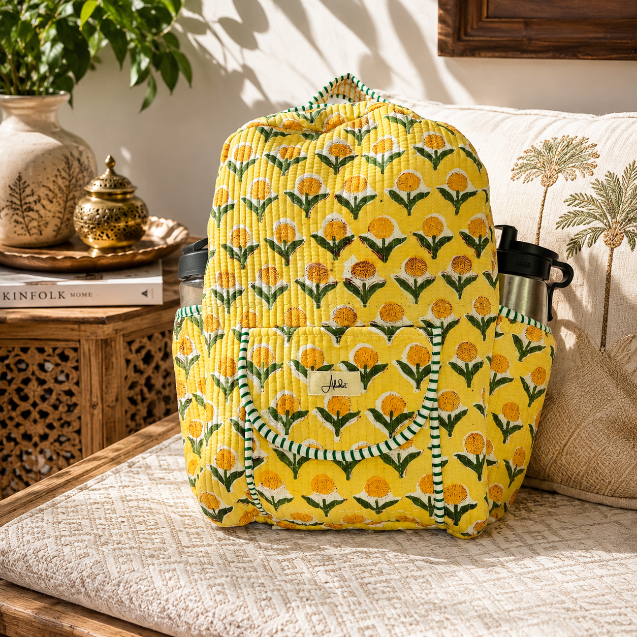 A yellow quilted backpack with a green and orange floral pattern, styled on a beige woven sofa next to a black water bottle.