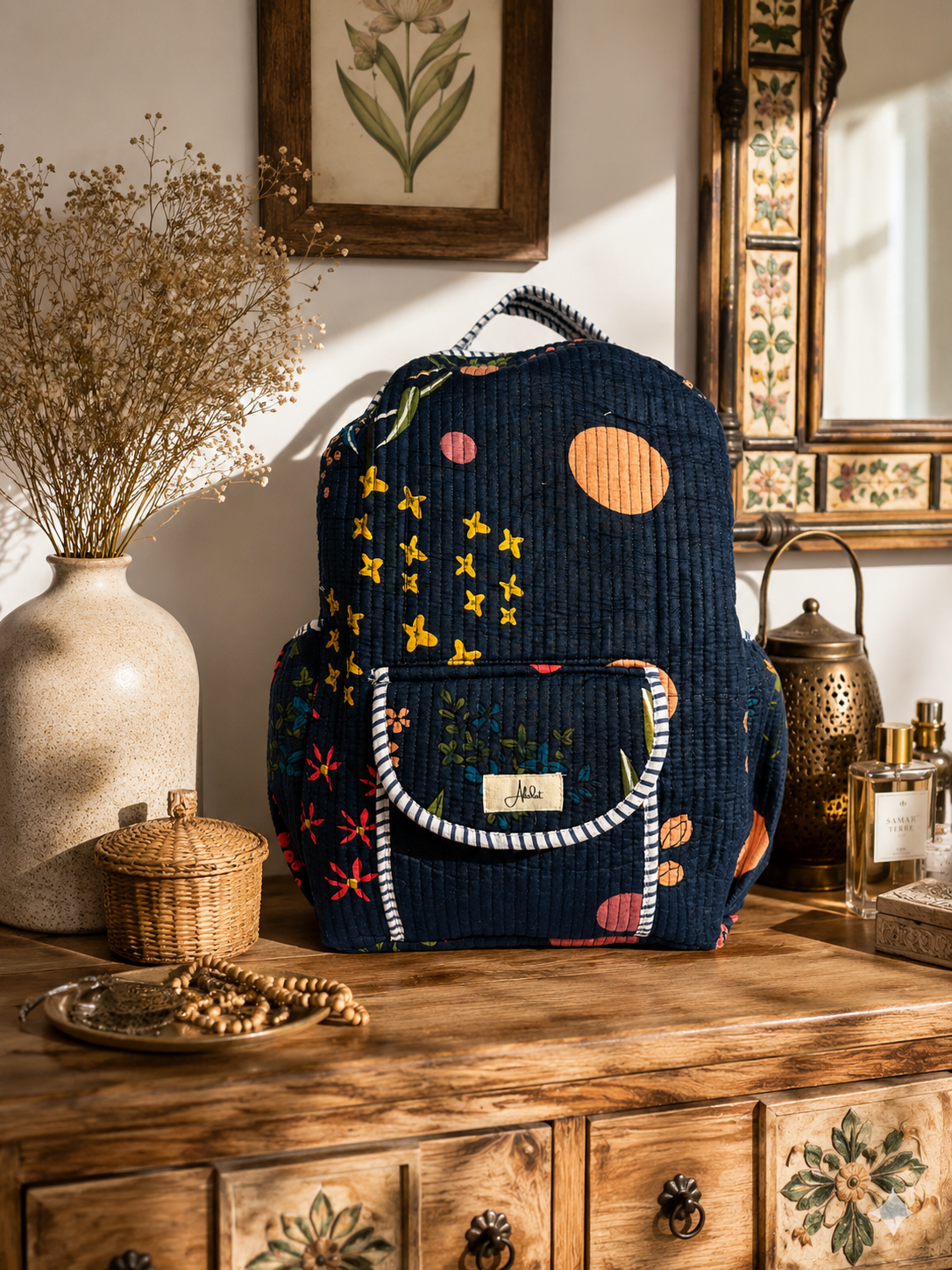 A navy blue quilted backpack with a colorful bohemian print of stars, circles, and floral motifs, displayed on a rustic wooden dresser in a warm, sunlit room.