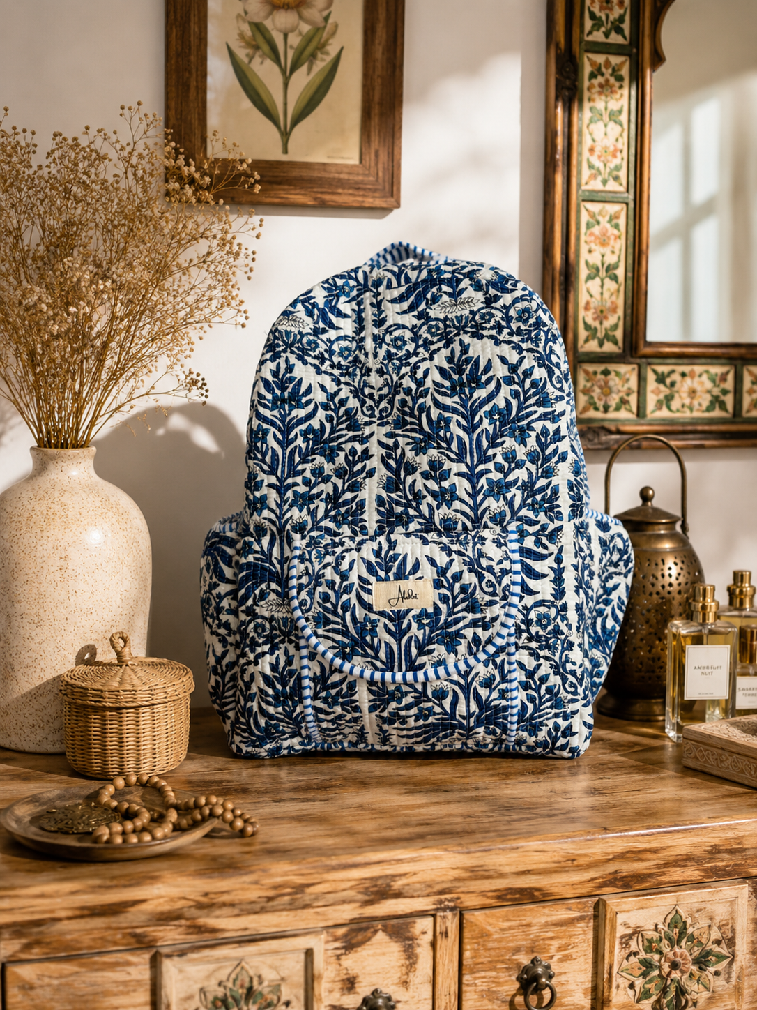 A blue and white patterned fabric quilted backpack styled on a rustic wooden chest in a warm, vintage-inspired room setting with a vase, dried flowers, and mirrors.