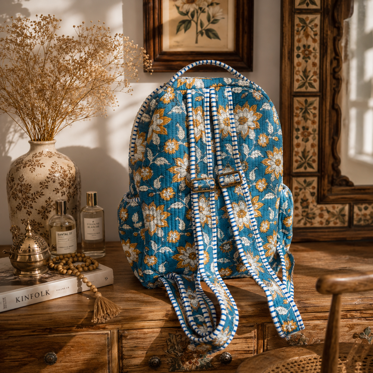 A blue floral backpack with white and blue patterned adjustable straps with metal adjusters, styled on a rustic wooden table next to a vase of dried flowers and decorative bottles.