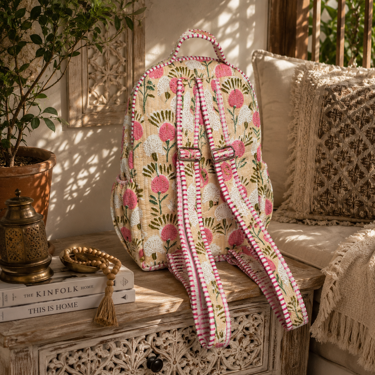 A floral backpack with pink and white pink and white stripey trim with adjustable straps with metal adjusters sits on a carved wooden table in a sunlit, bohemian-style room next to a couch with patterned pillows.
