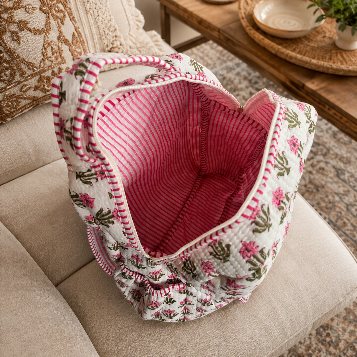 A quilted fabric tote bag with a pink and green floral pattern on the exterior and a pink striped lining, shown open on a beige upholstered chair in a living room setting.
