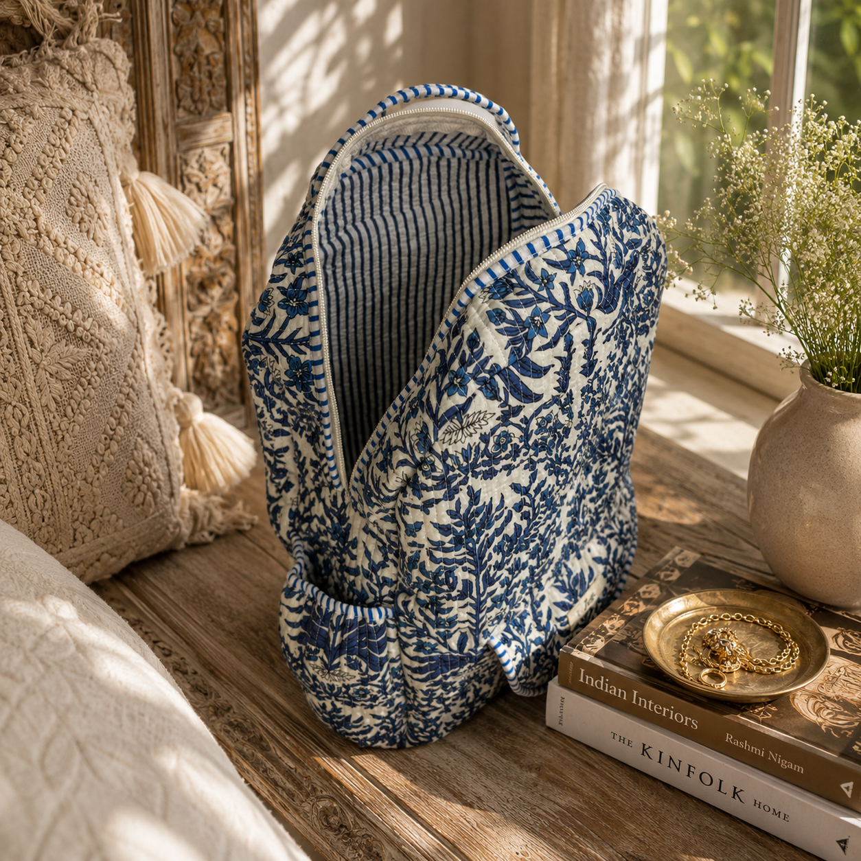 A blue and white floral patterned fabric backpack with blue and white striped interior stands open on a wooden surface in a sunlit room, styled beside a woven pillow, books, and a vase with dried flowers.