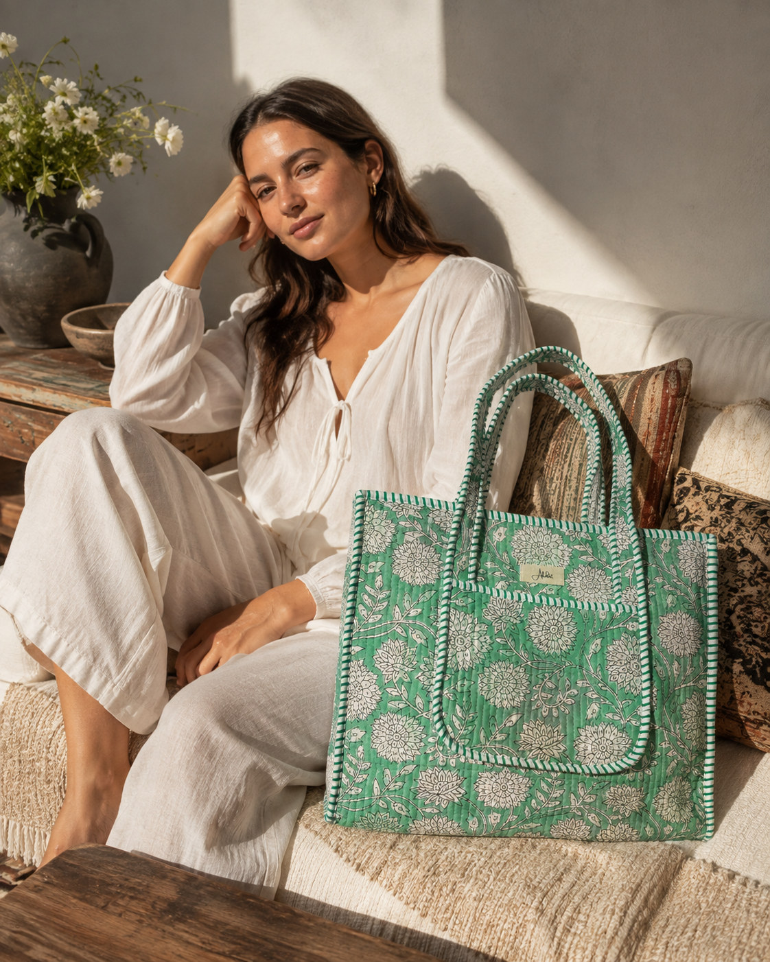A woman in a white lounge set sits on a sunlit sofa next to a green quilted tote bag with white floral blockprint patterns and green-and-white striped handles.