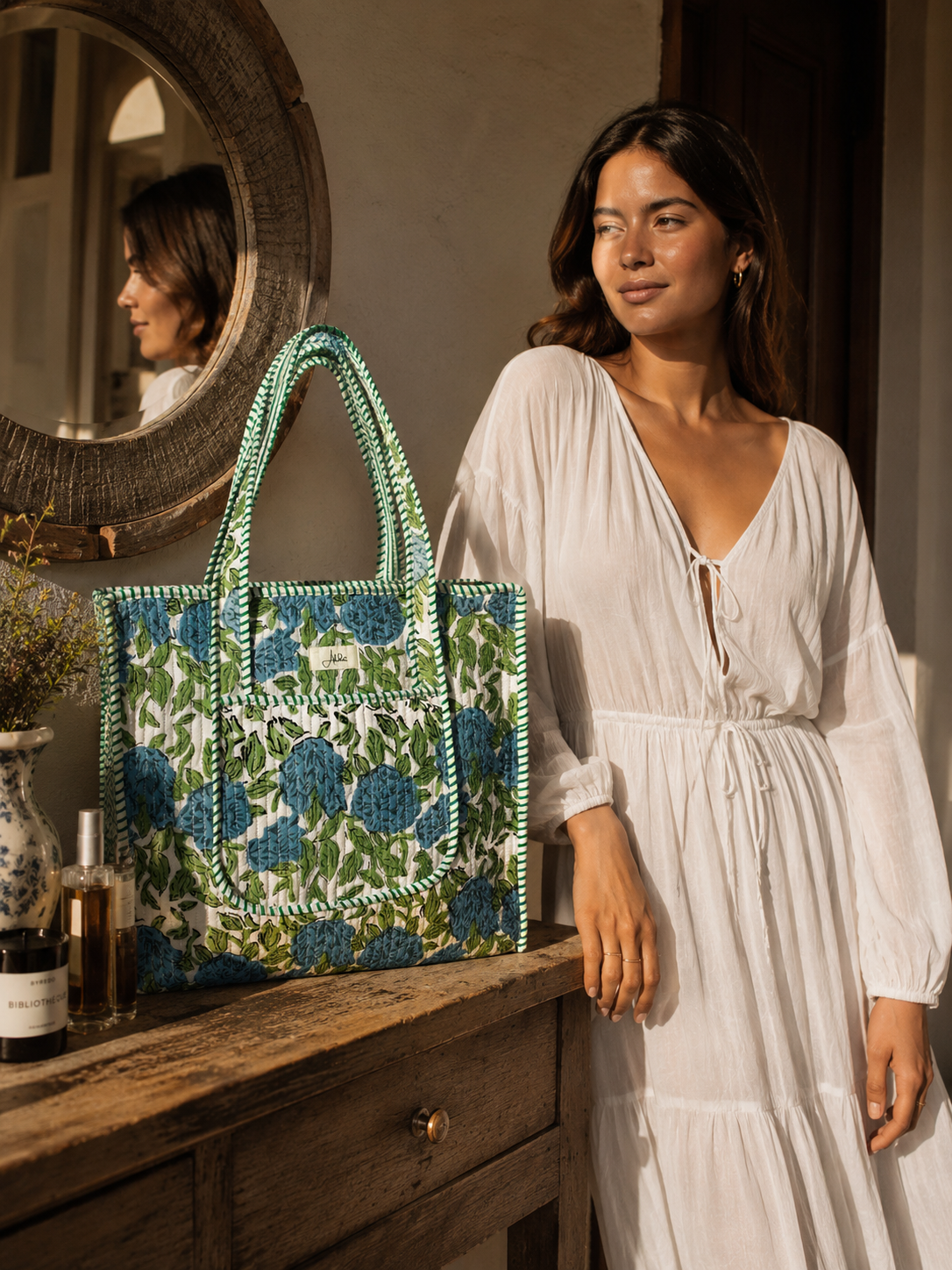 A woman in a white dress stands beside a rustic wooden table, holding a large patterned quilted tote bag with blue flowers and green leaves. A round mirror on the left reflects her profile, and bottles and a vase sit on the table.