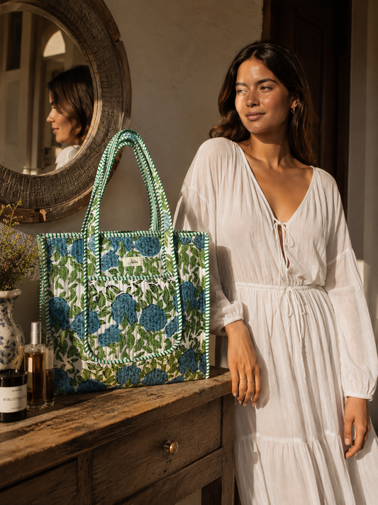 A woman in a white dress stands beside a rustic wooden table, holding a large patterned quilted tote bag with blue flowers and green leaves. A round mirror on the left reflects her profile, and bottles and a vase sit on the table.