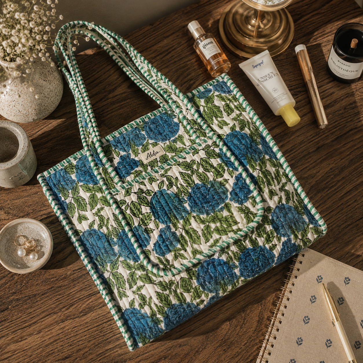 A quilted tote bag with a blue floral pattern and green leafy trim, styled on a dark wooden table with lifestyle props including skincare items, a candle, and stationery.