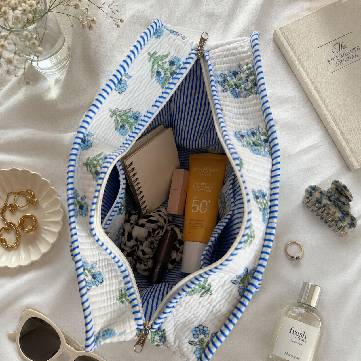 A quilted blockprint tote bag with a blue and white striped border and a blue and white striped interior, shown open on a white fabric background with various lifestyle accessories and cosmetics inside. The bag has 3 pockets inside. 