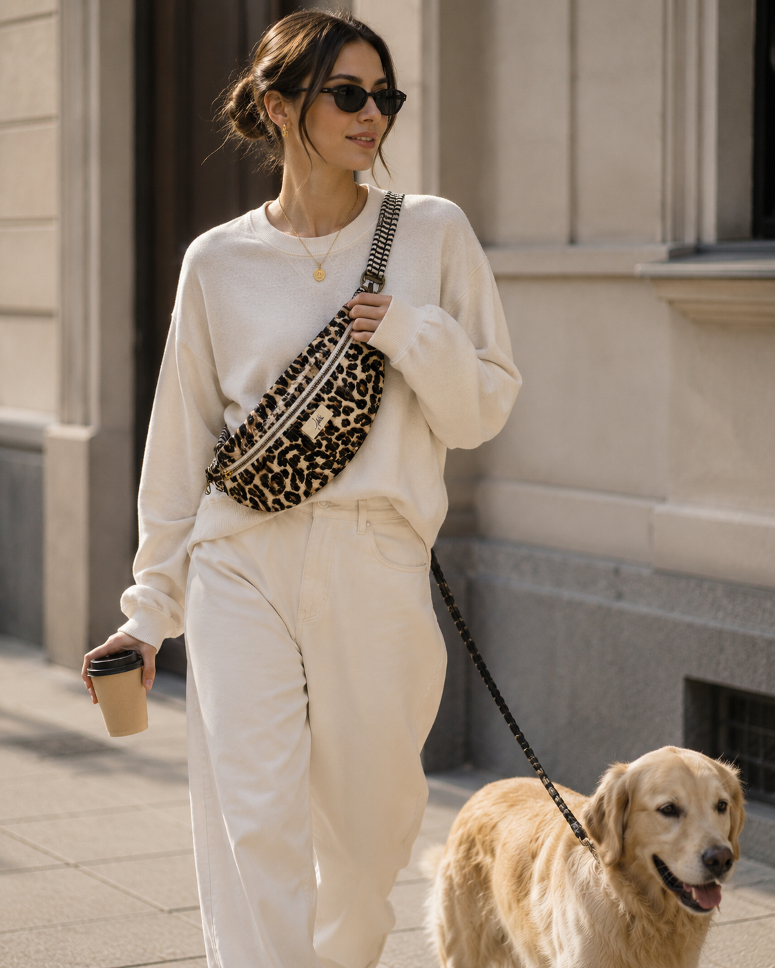 A woman wearing a cream sweatshirt and matching sweatpants walks with her golden retriever dog, carrying a leopard-print crossbody bag and holding a coffee cup in an outdoor urban setting.