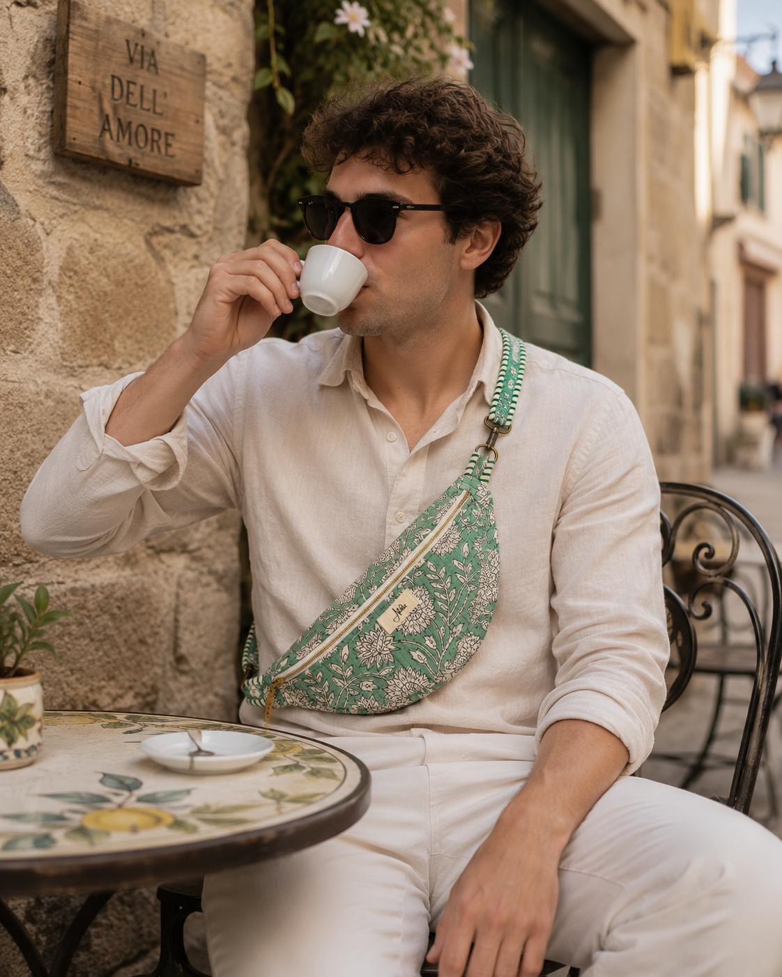 A man wearing a green and white patterned fabric crossbody fanny pack sits at an outdoor cafe table, drinking from a white cup.