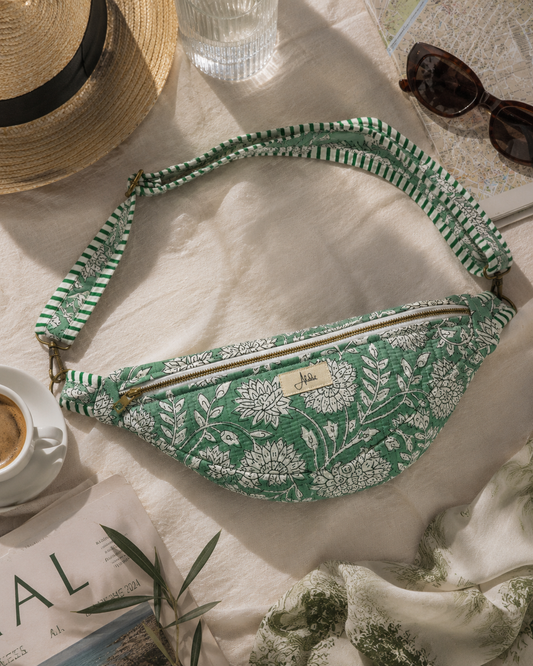 A green and white floral print fanny pack with a matching patterned strap, styled in a flat-lay arrangement with a straw hat, sunglasses, a coffee cup, and a map on a beige fabric background.