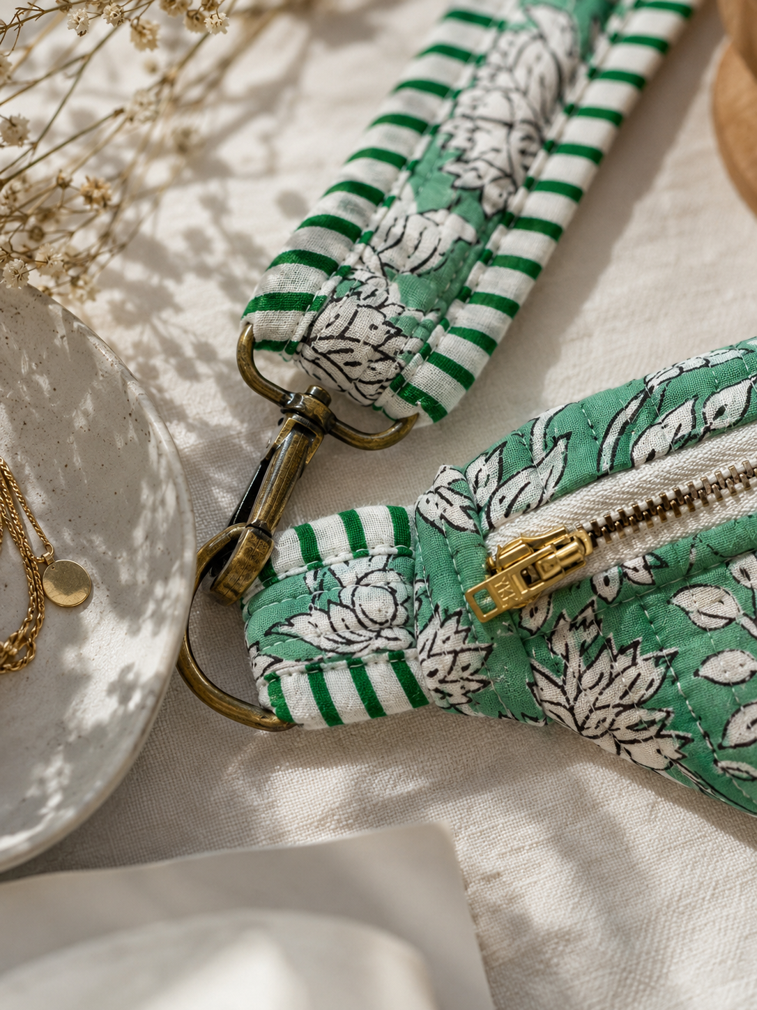 A close-up of a fabric zipper pouch with a green and white floral print and striped trim, attached to a matching wrist strap with brass-tone hardware, styled on a light fabric surface with dried flowers and a plate.