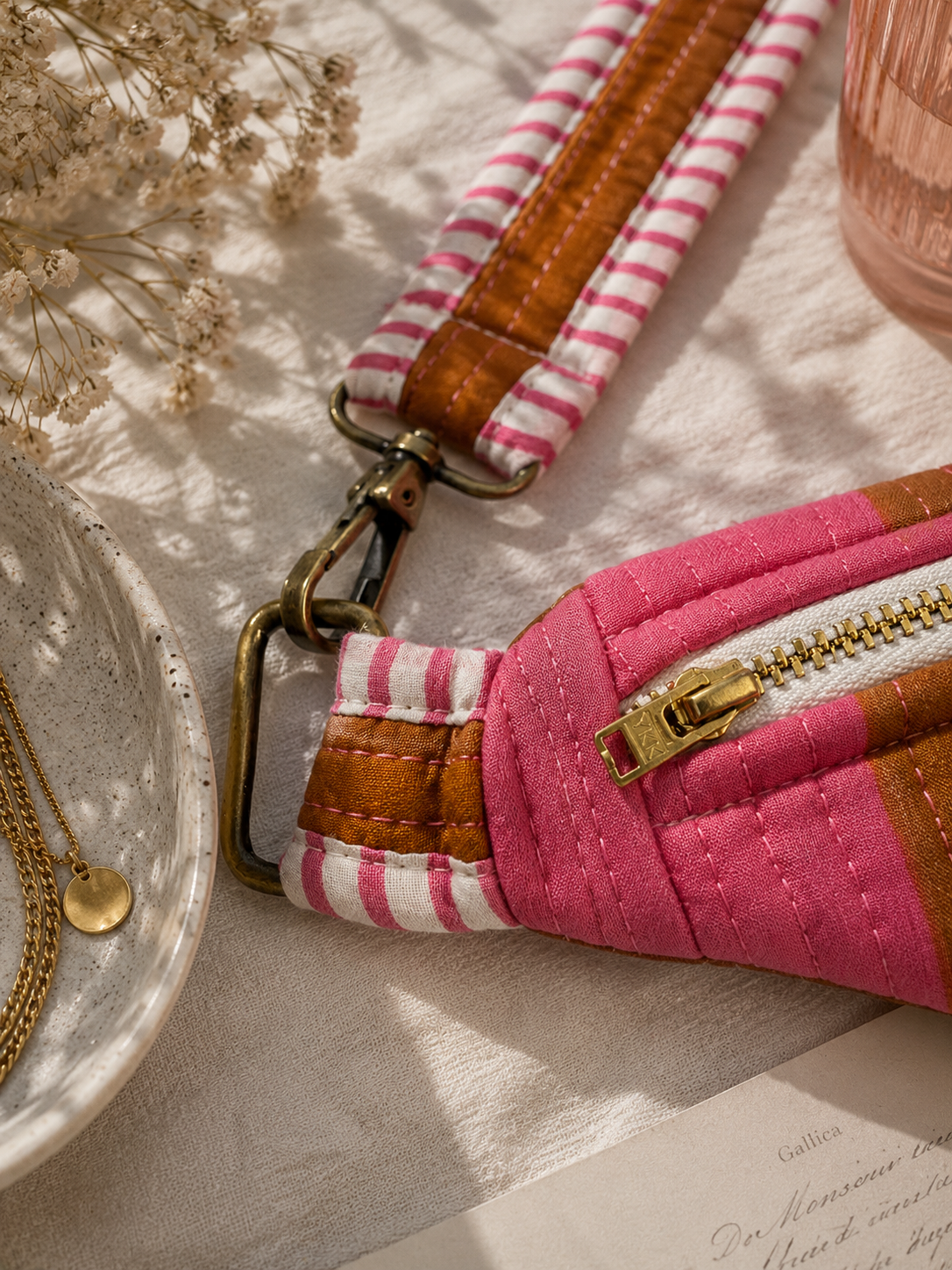 A close-up of a pink and rust-stripped quilted crossbody bag with a pink and white striped fabric strap, brown leather trim, and gold-tone hardware, styled on a textured cream fabric with dried flowers and a ceramic dish.