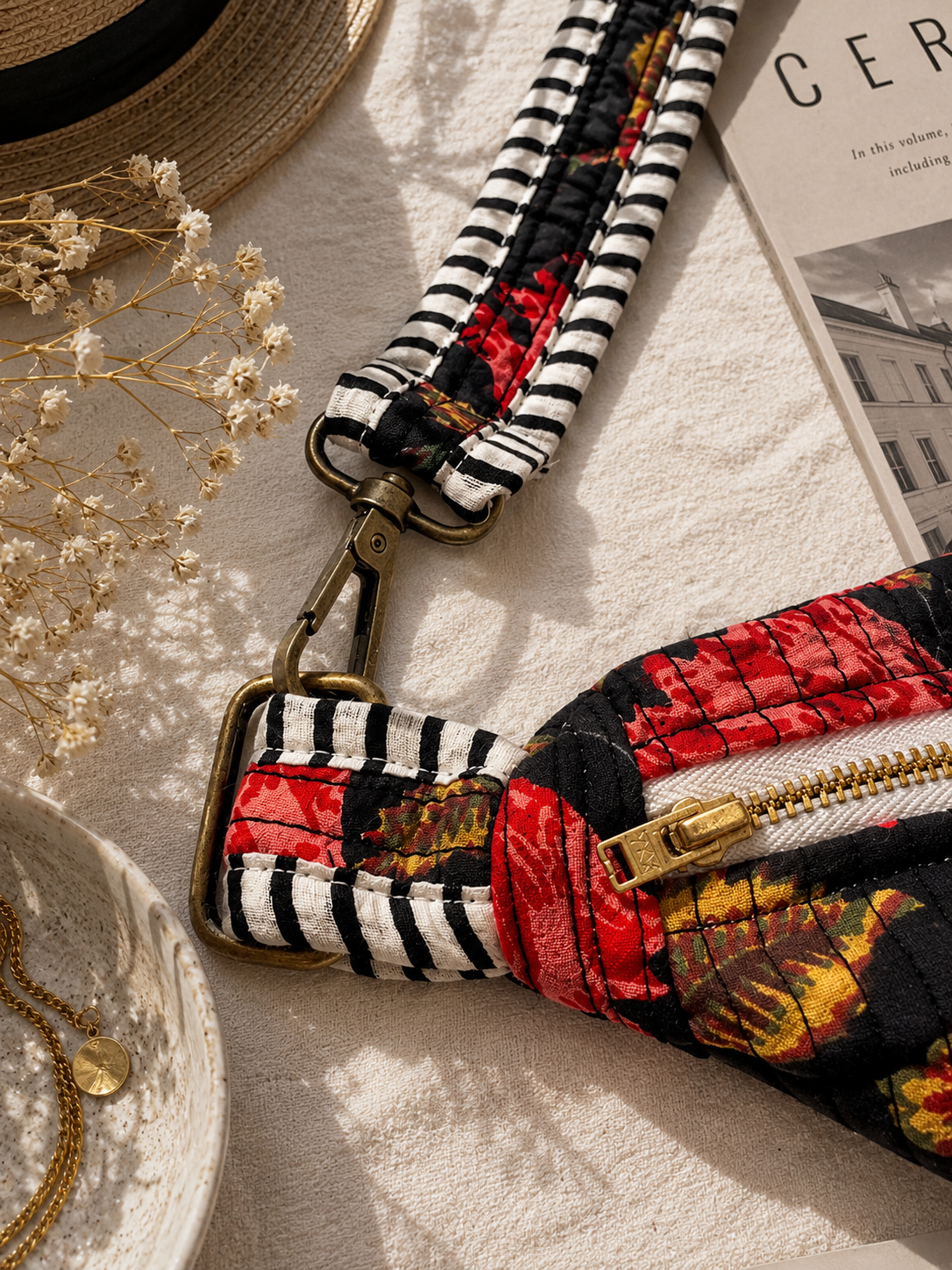 A close-up of a fabric crossbody bag strap and zipper section laid on a light textured surface with styled props including dried flowers, a hat, and a magazine. The bag has a black-and-white striped woven strap, a red and black floral print body, and gold-tone hardware.