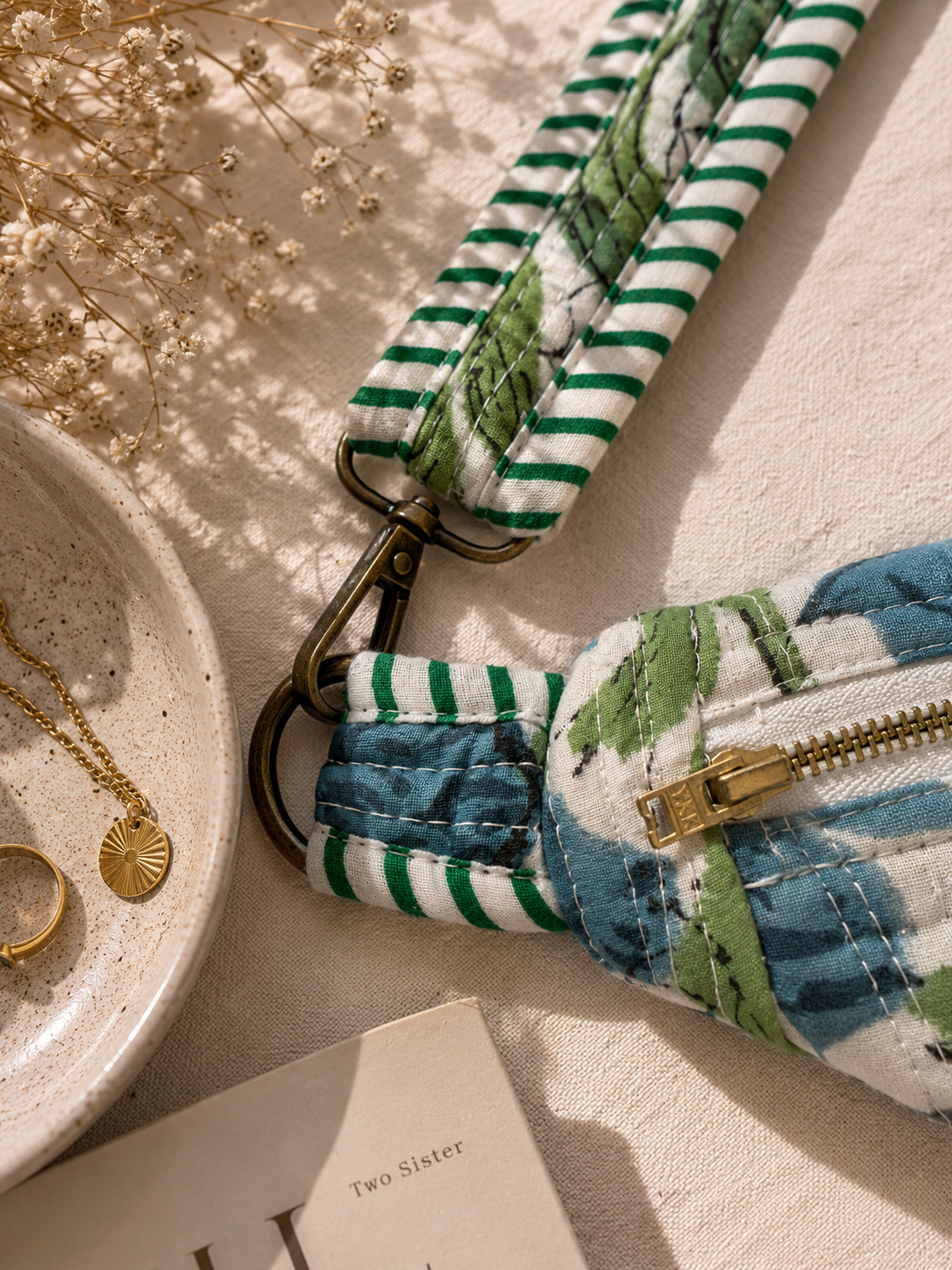 A close-up of a fabric wristlet strap with a green and white striped section and a blue and green floral print section, attached to a metal clasp on a beige surface with styled props.