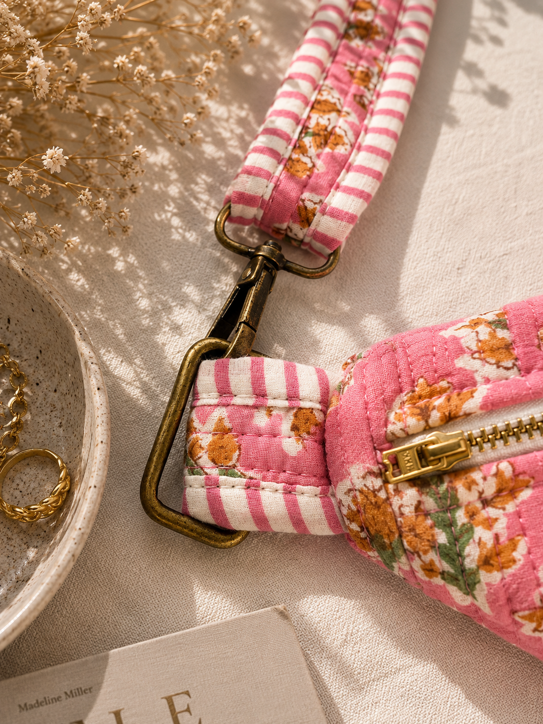 A close-up of a pink and white striped fabric strap with a floral patterned pouch and antique brass hardware, styled on a textured beige fabric with dried flowers and a ceramic dish.