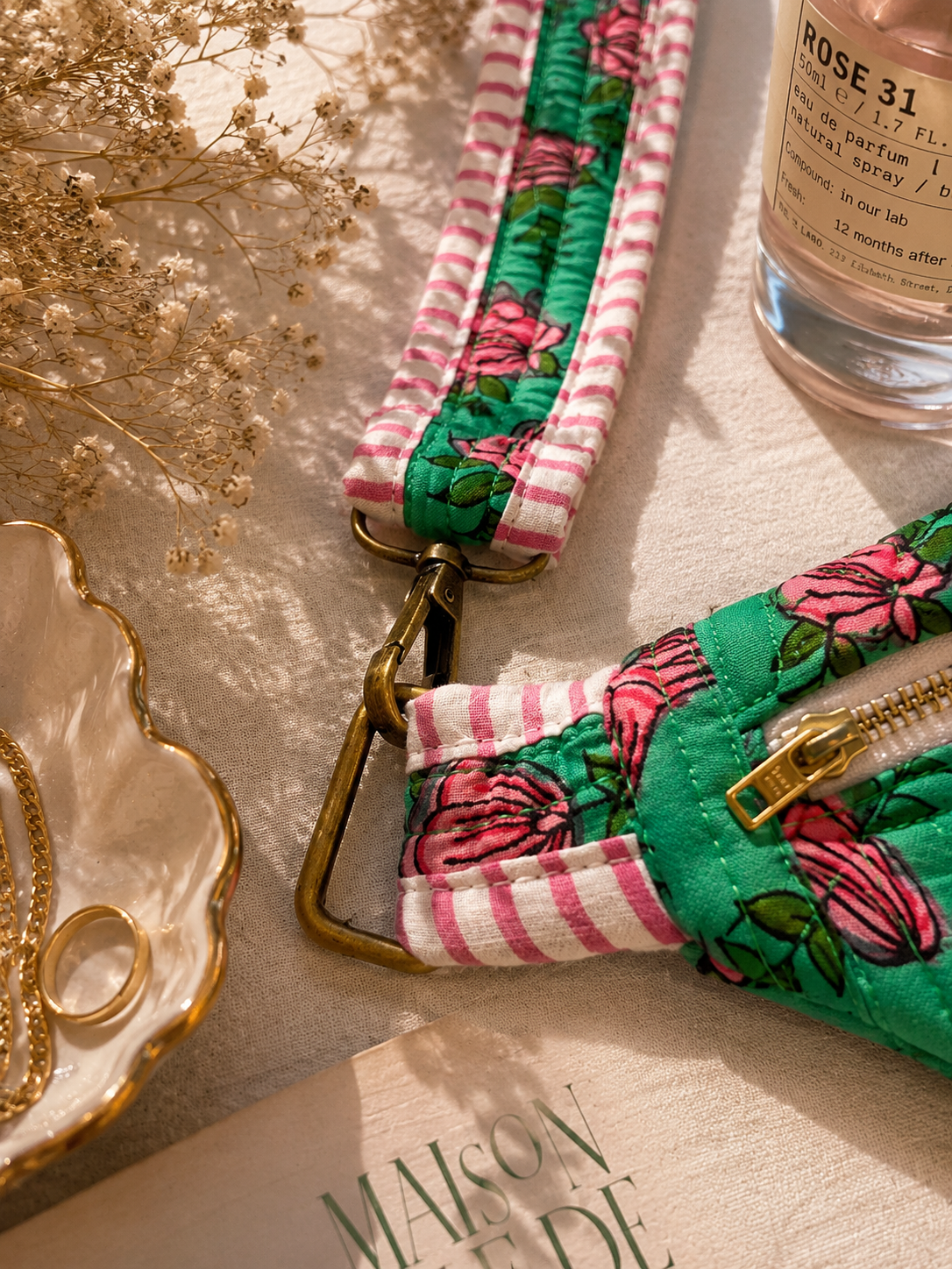 A close-up styled flat-lay of a quilted fabric strap or pouch with a green floral print, pink-and-white striped fabric, and brass-tone hardware, arranged on a beige surface with dried flowers, rings, and a perfume bottle nearby.