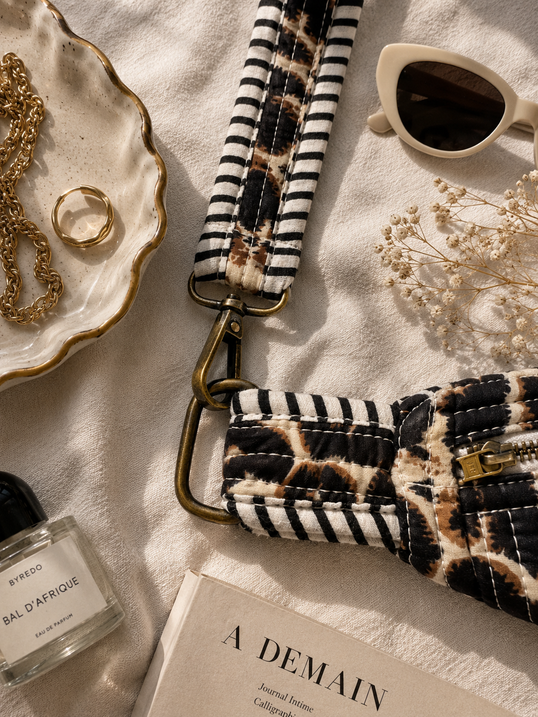 A close-up flat lay of a fabric shoulder strap bag with a black-and-white striped section and a black-and-brown leopard print section, attached to a gold-tone clasp. The bag is styled on a light fabric surface with sunglasses, jewellery, a perfume bottle, dried flowers, and a book.