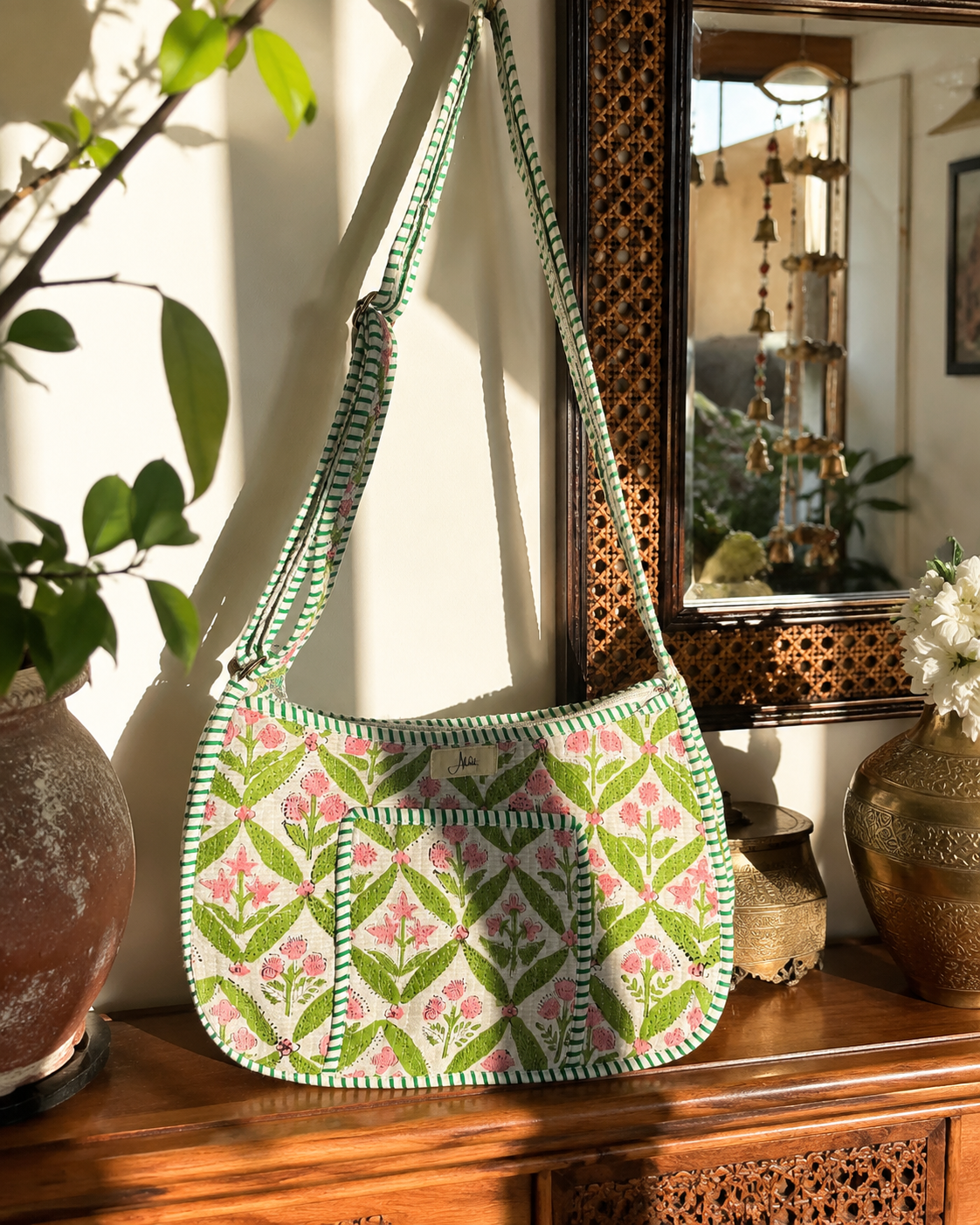 A green and pink floral-patterned shoulder bag with a matching adjustable strap, displayed on a wooden cabinet in a sunlit room with decorative plants and a mirror.