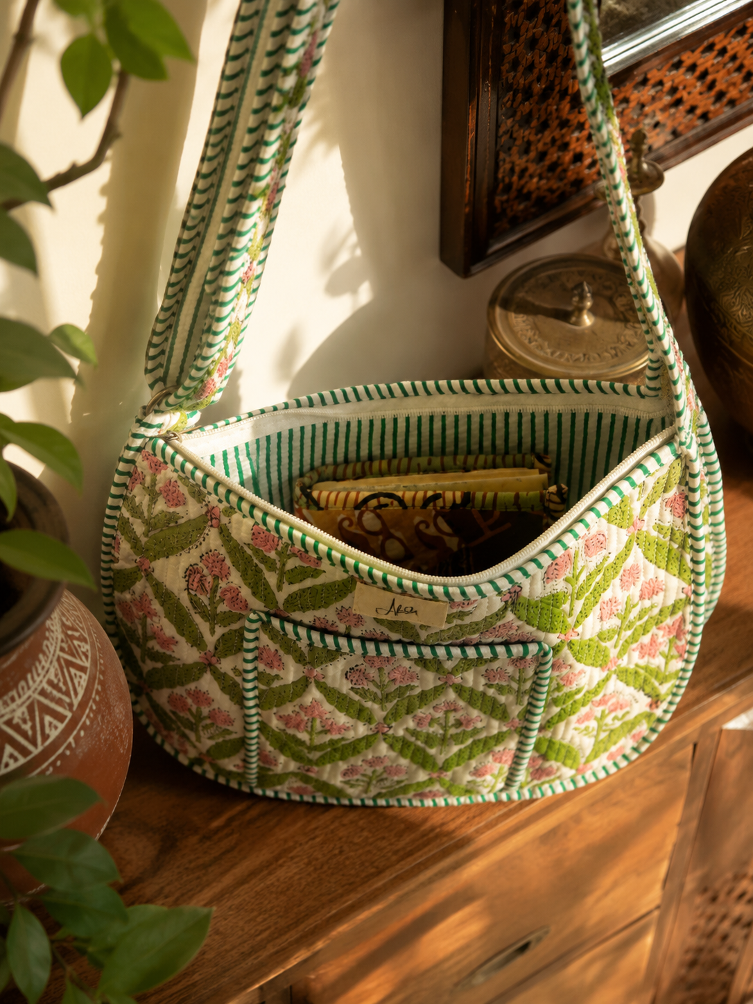 A quilted shoulder bag with a green and pink floral pattern and striped trim, resting on a wooden table in a warm, sunlit room.