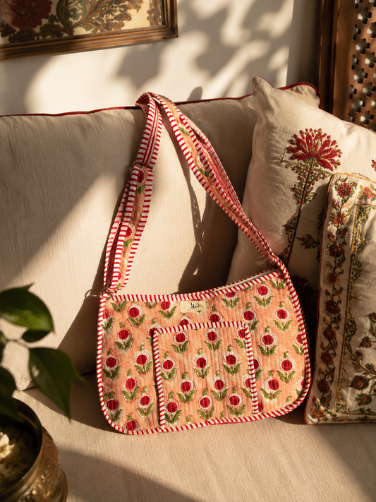 A peach-coloured embroidered quilted shoulder bag with red and green floral patterns and red-and-white striped trim, resting on a beige sofa with decorative pillows in a sunlit room.